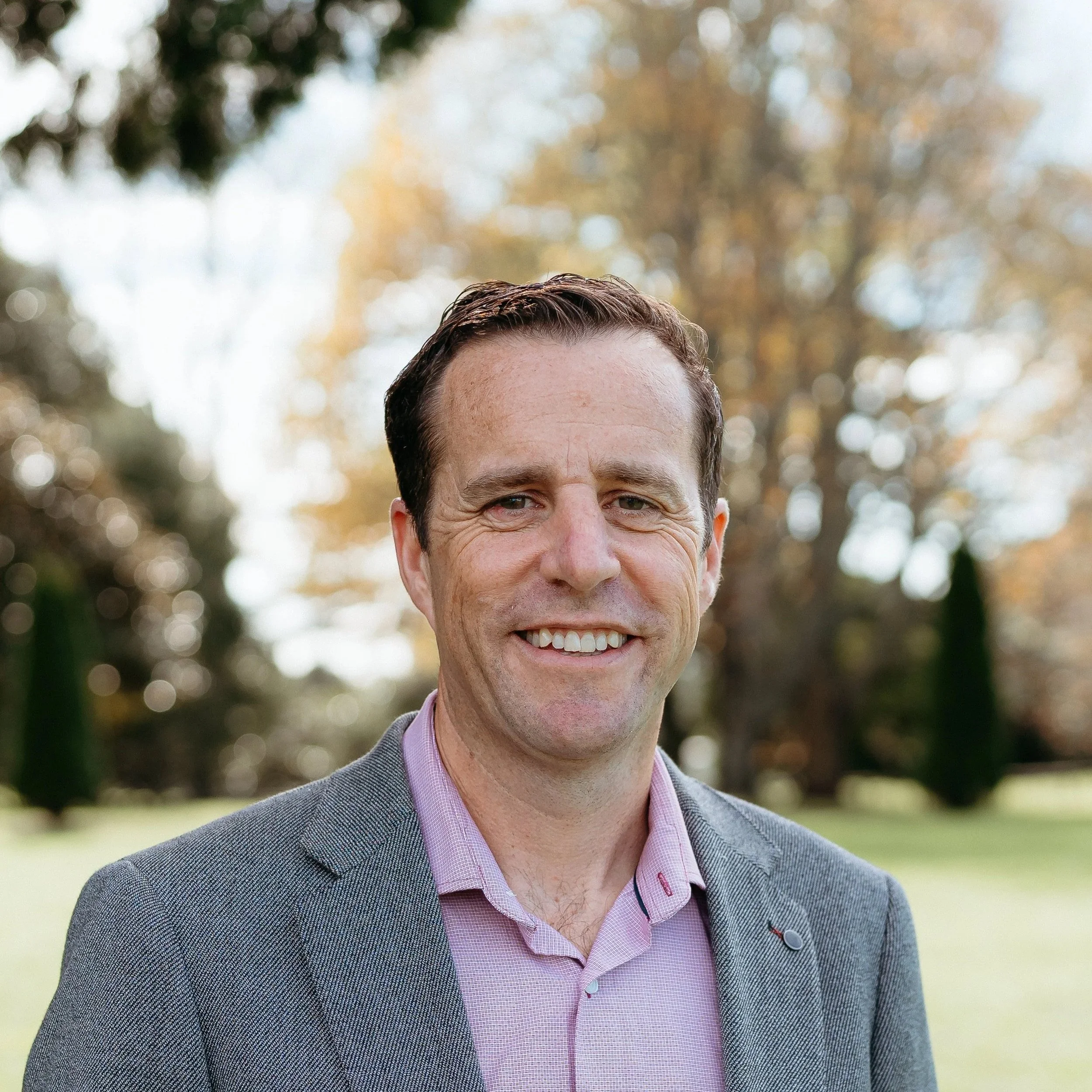 Portrait of a smiling man wearing a gray blazer and pink shirt outdoors with trees and foliage in the background