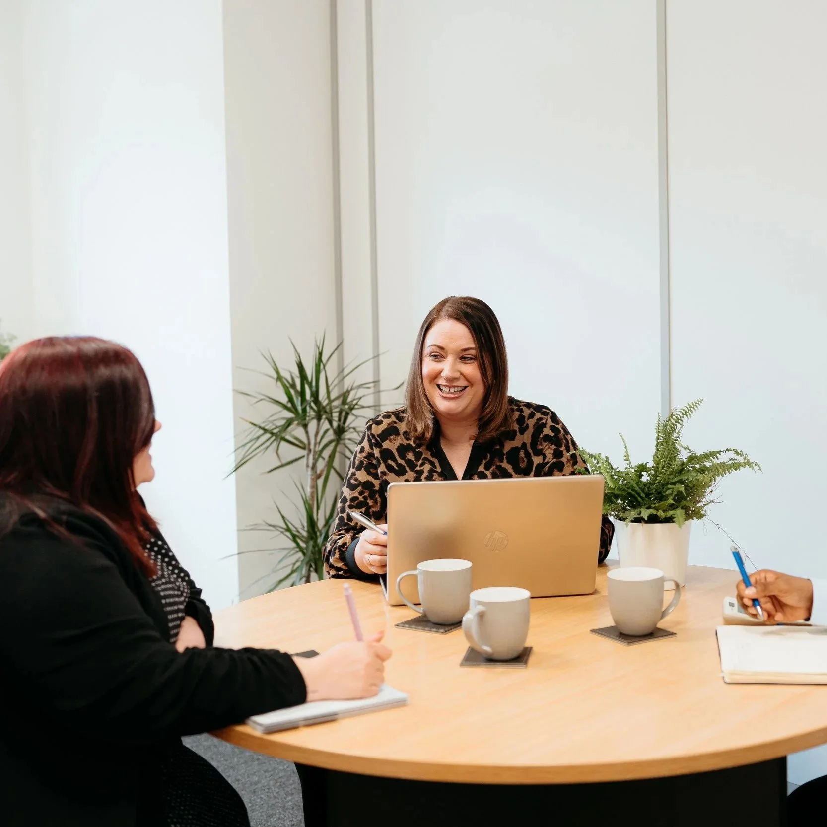 Three women having a meeting around a wooden table in an office, one woman is speaking and smiling, with laptops, notebooks, and coffee mugs on the table, and a potted plant in the background.