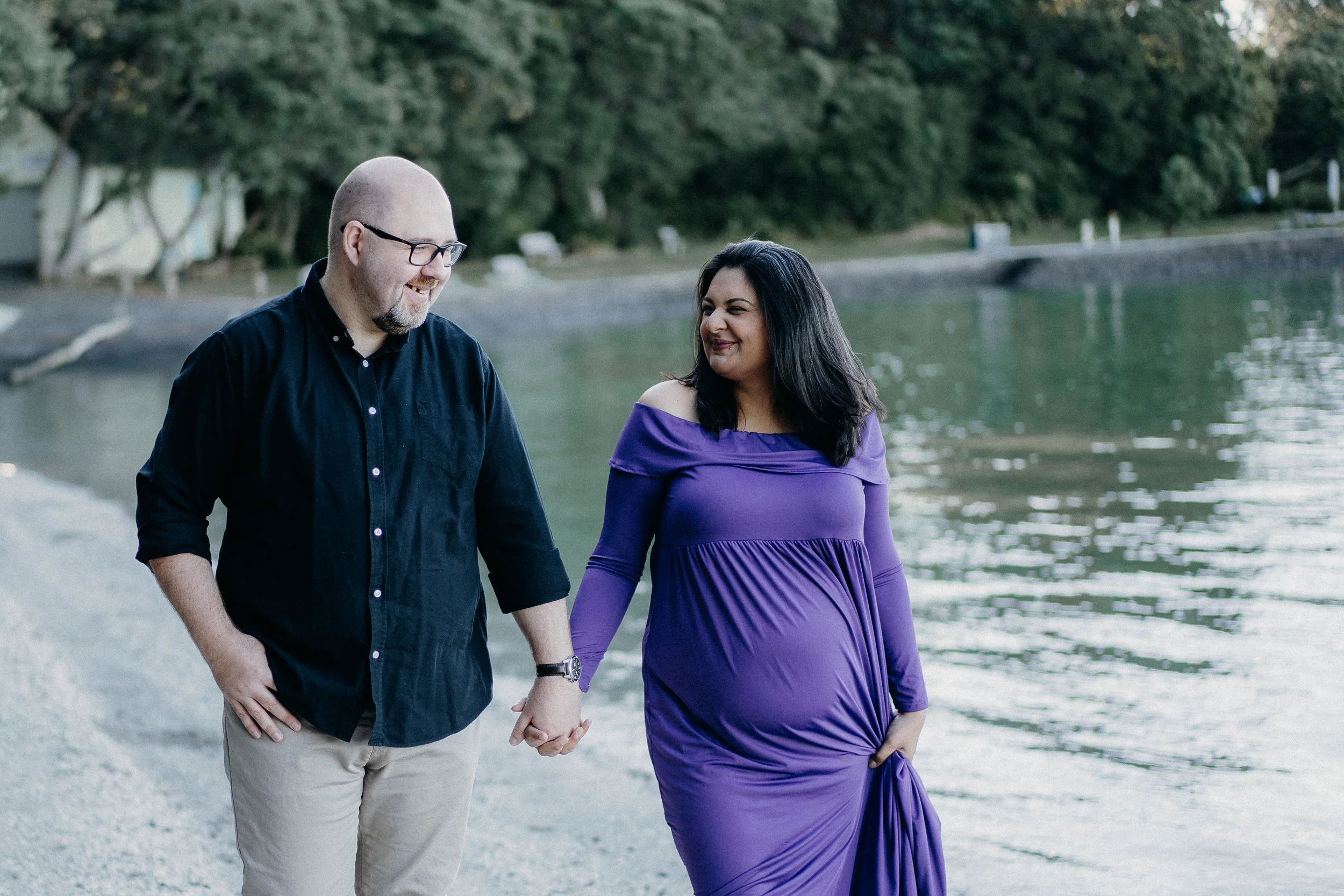 A diverse couple holding hands and walking along a lakeshore, with trees and water in the background during daytime.