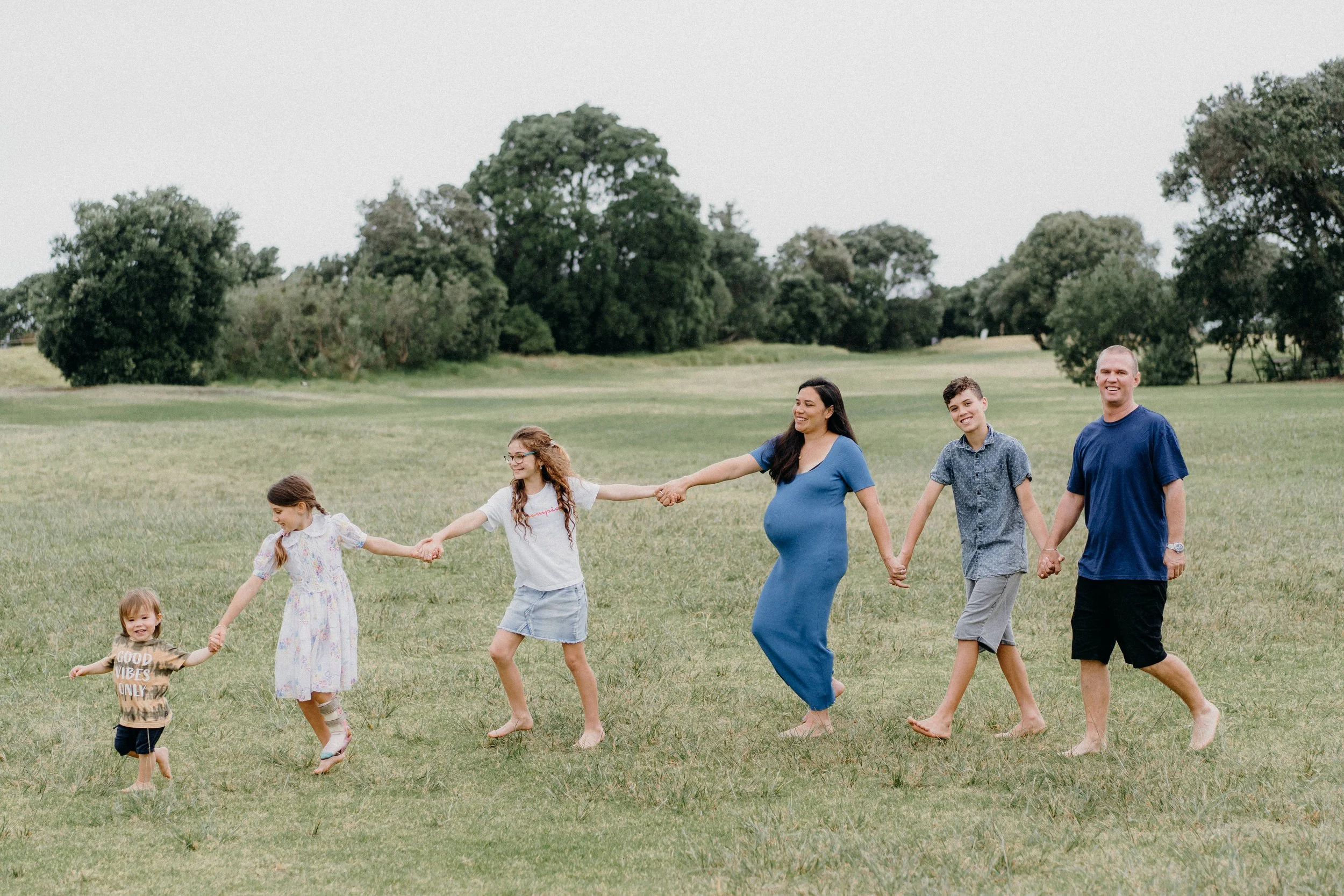 Family holding hands and walking across a grassy field outdoors, including adults and children of various ages.