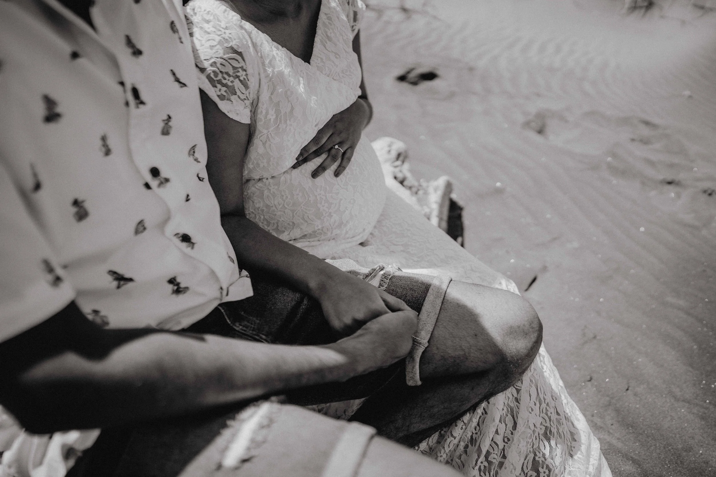 A black and white photo of a couple sitting on a bench at the beach, with the woman showing her pregnant belly and the man holding a wide-brimmed hat in his lap.