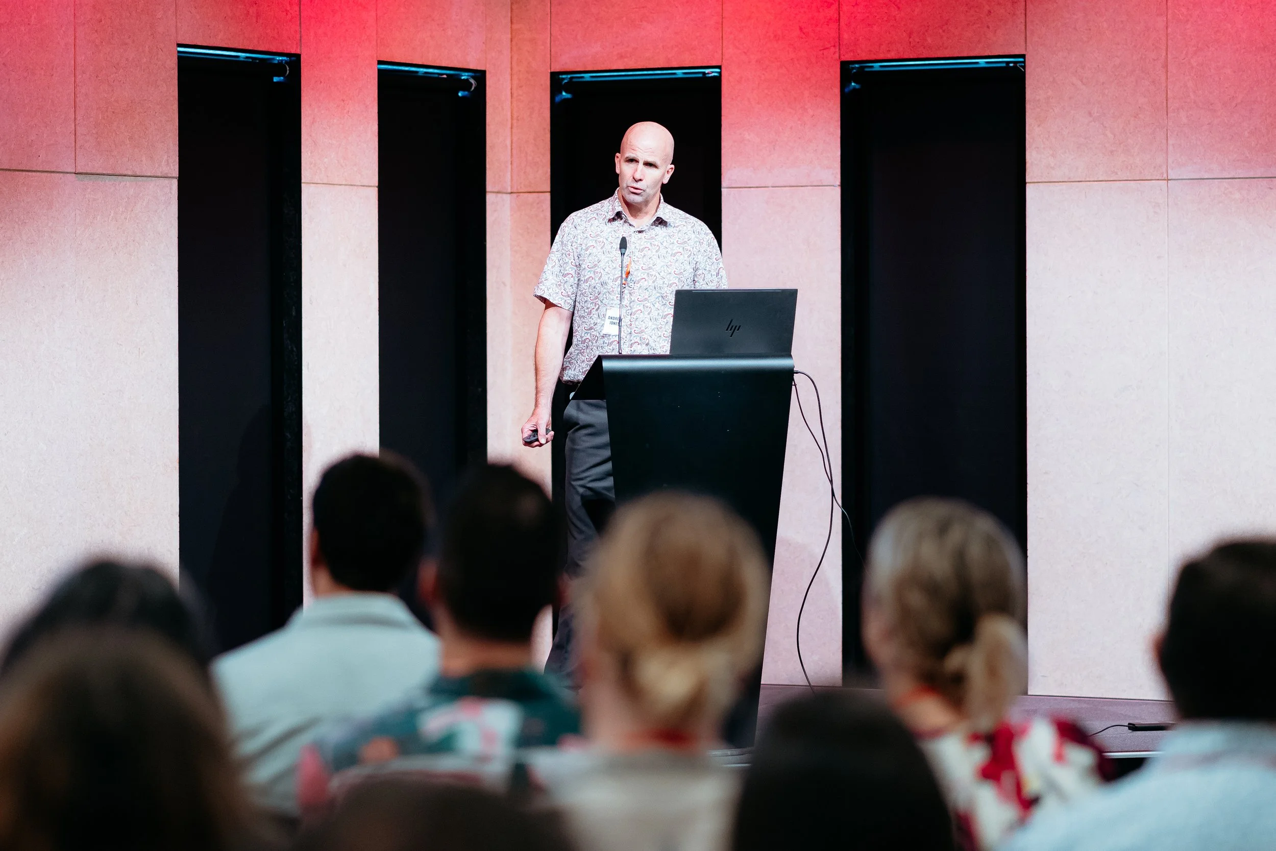 A man giving a presentation at a conference, standing behind a podium with a laptop, audience in foreground.