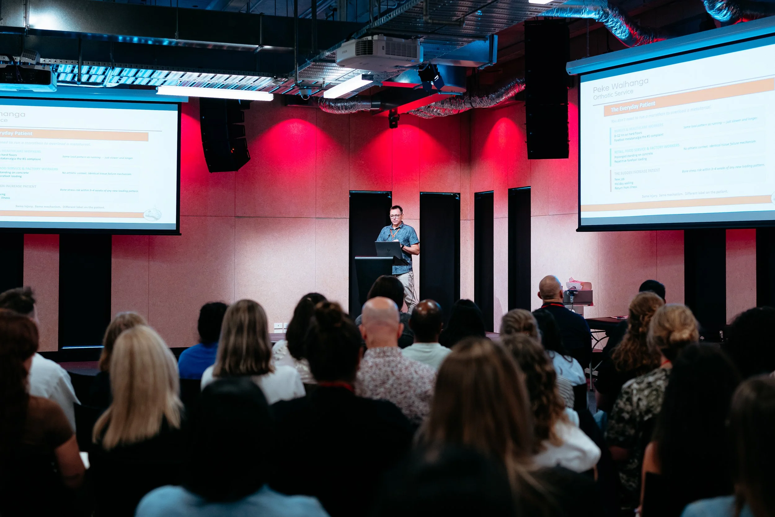 A speaker presents at a conference in front of a seated audience, with two large screens displaying a slide titled 'Peke Waihanga Orthopedic Service'.