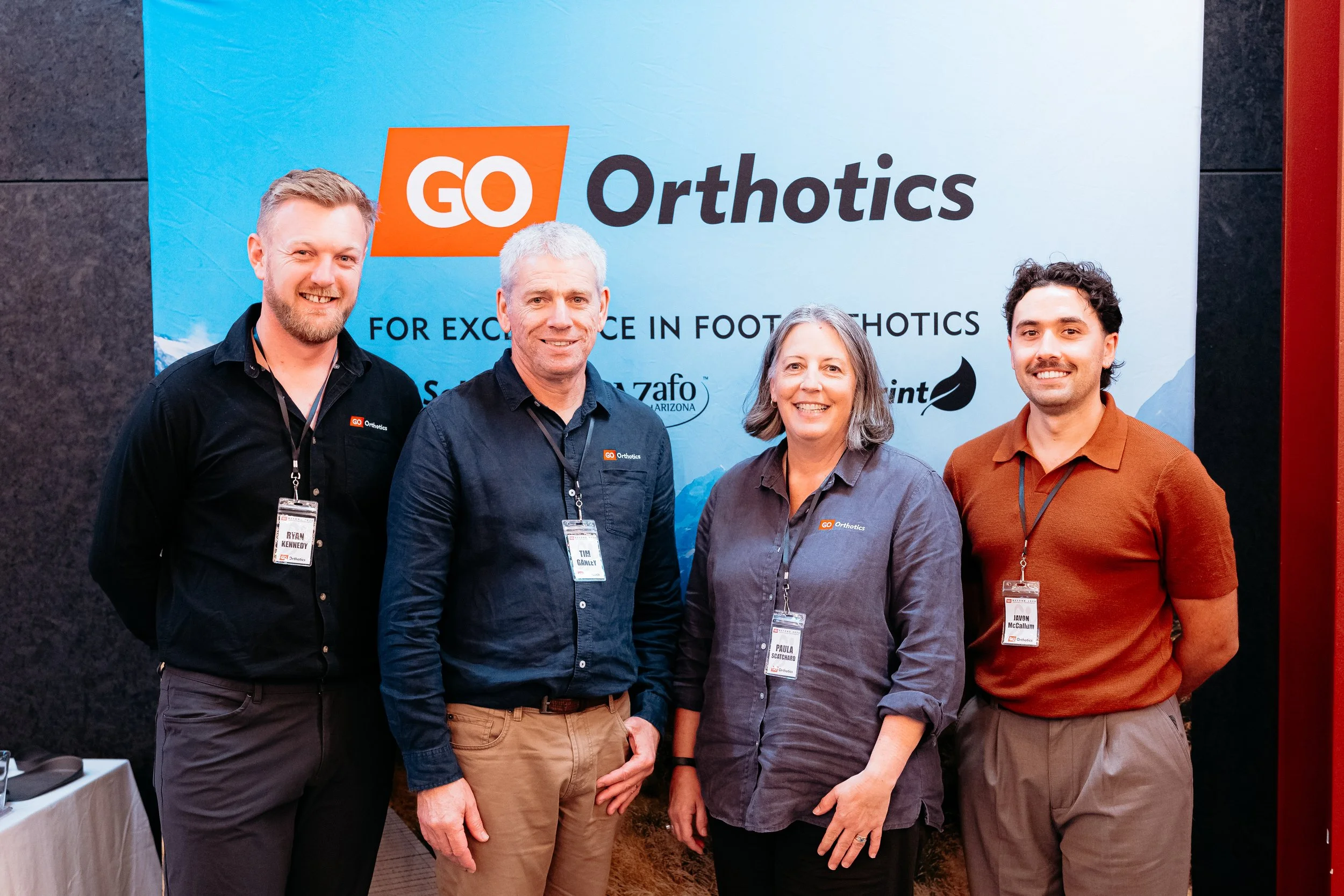 Four people standing indoors in front of a large GO Orthotics banner, wearing name tags and business casual attire.