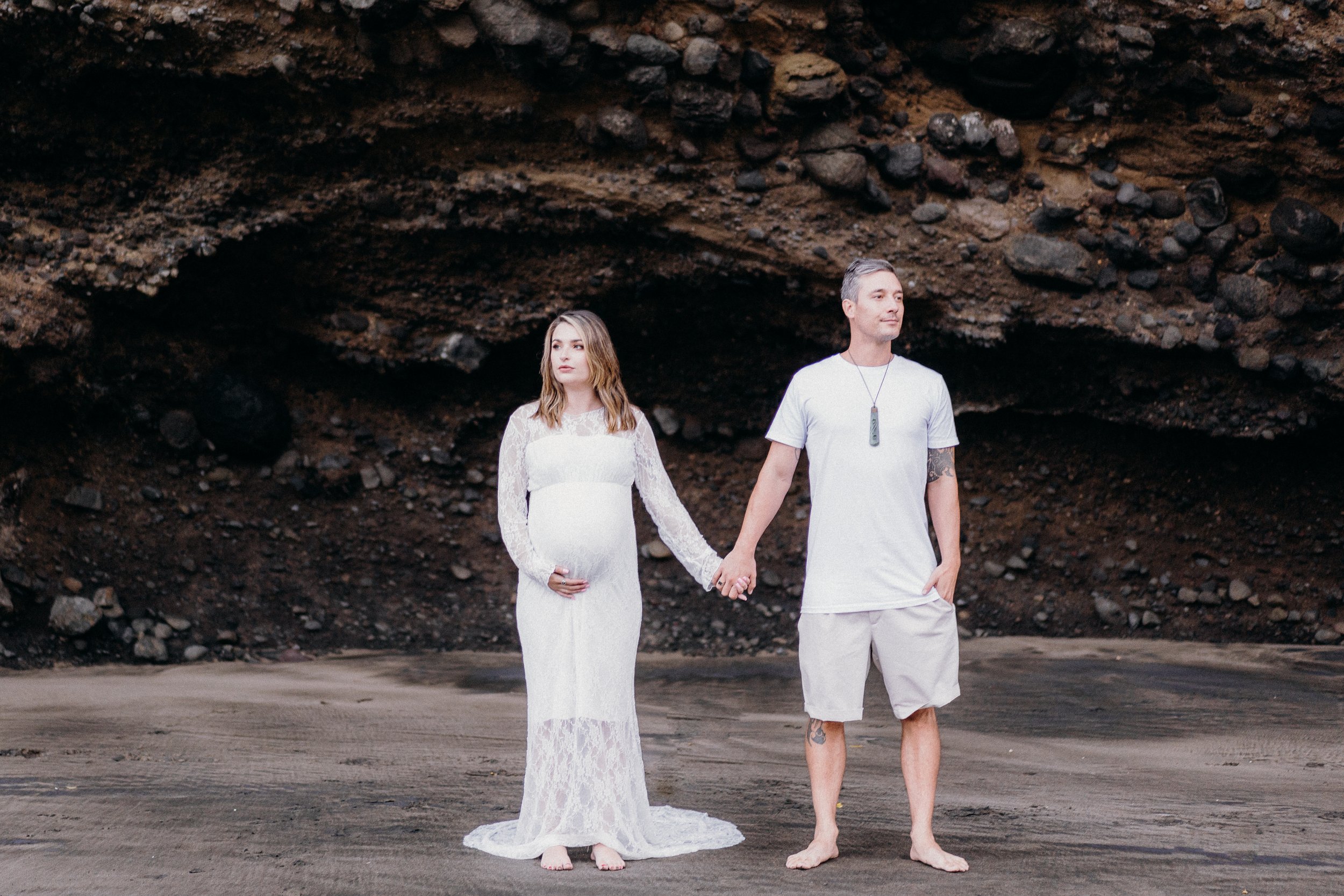 Pregnant woman in a white lace dress holding hands with a man in white shirt and beige shorts, standing barefoot on a rocky beach.