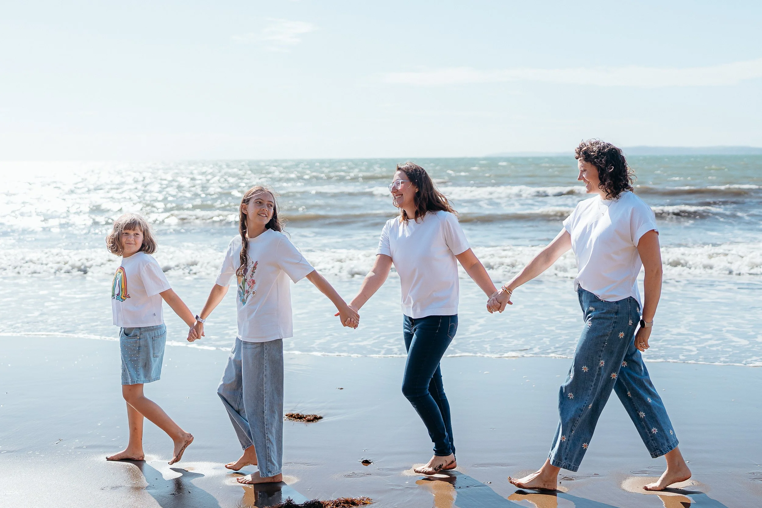 A happy family of four, including a man, woman, a young girl, and a baby, enjoying a moment together on a sandy beach with rocky cliffs and trees in the background, smiling and looking at each other.
