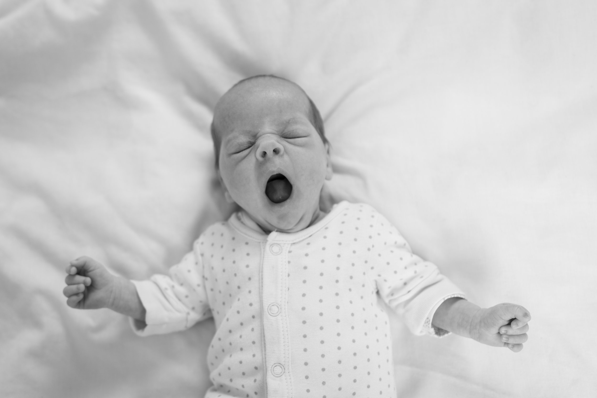 Black and white photo of a yawning baby in a polka dot onesie lying on a white blanket.