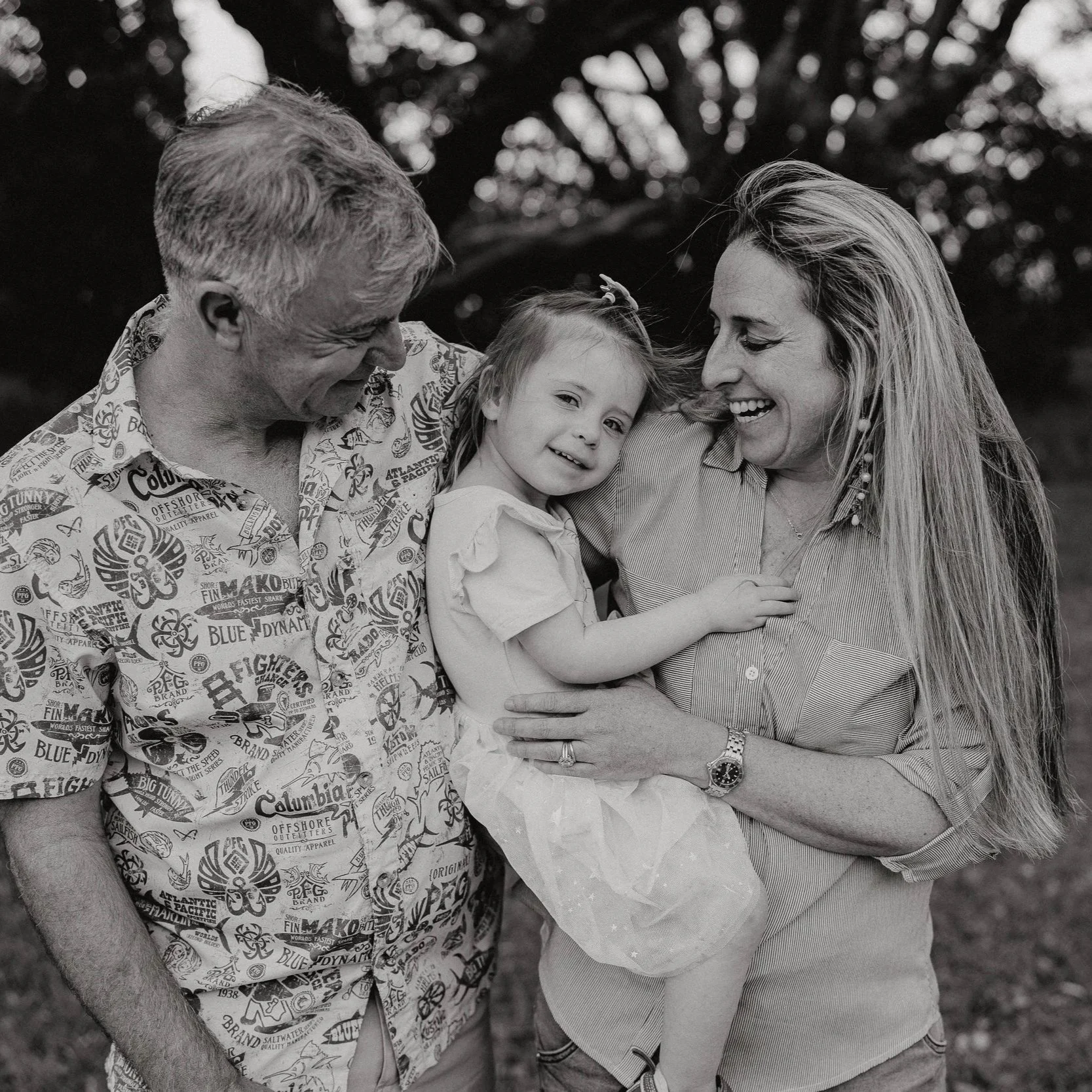 A black and white photo of a man, woman, and young girl outdoors. The man and woman are smiling and looking at each other, while the girl is leaning on the woman and smiling at the camera.