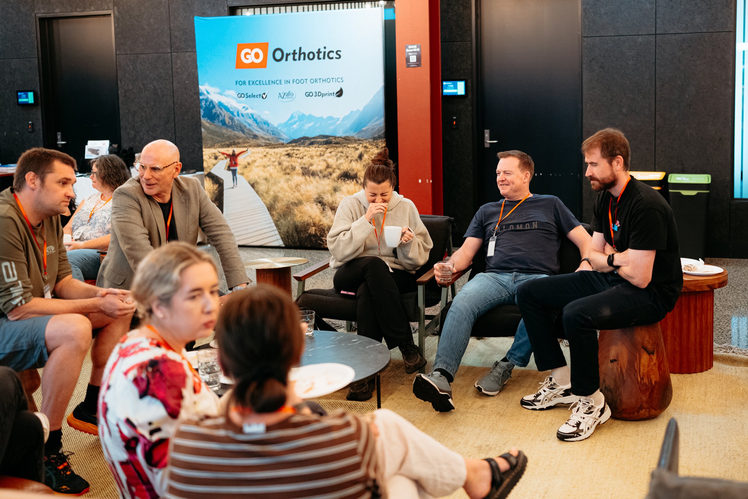 Group of people sitting and chatting in a room with a large poster for GO Orthotics in the background.