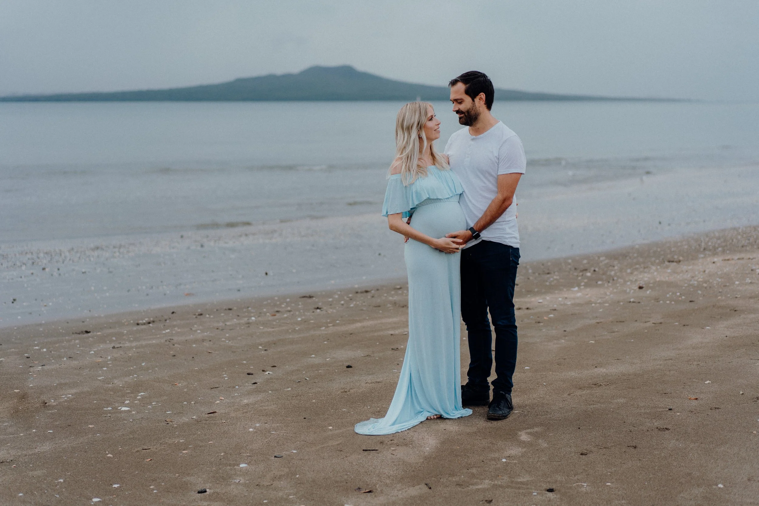 A pregnant woman and a man standing on a beach, holding her belly, gazing at each other with a mountain in the background.
