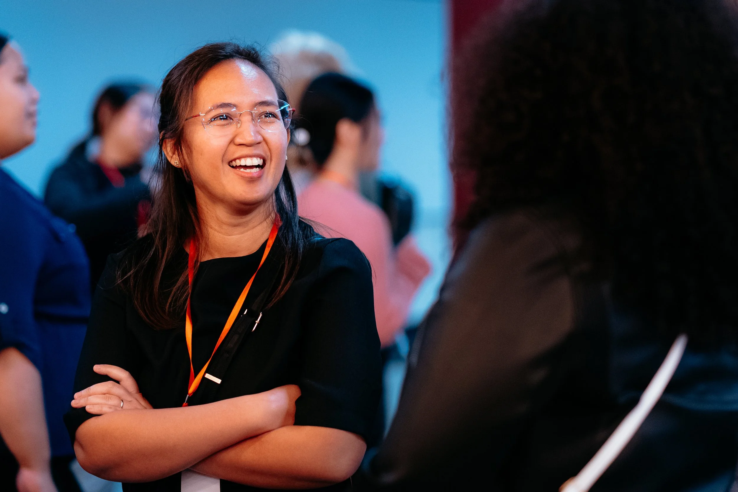 A woman with glasses and long dark hair smiling and talking with others at a crowded indoor event.