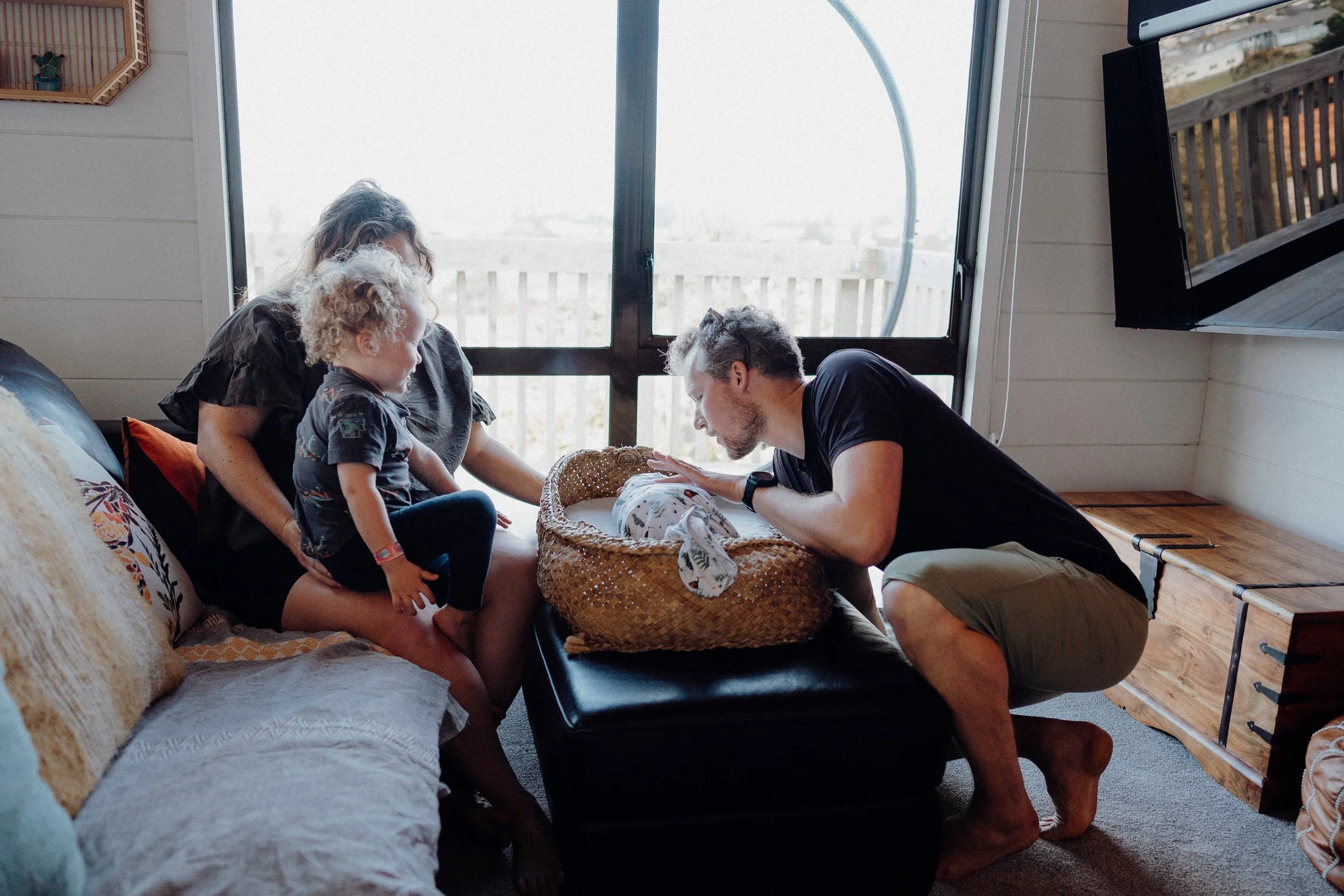 A family gathered around a newborn baby in a wicker bassinet, with a woman and a young child closely observing, and a man kneeling and gently touching the baby, inside a cozy room with a sliding glass door and a wall-mounted television.