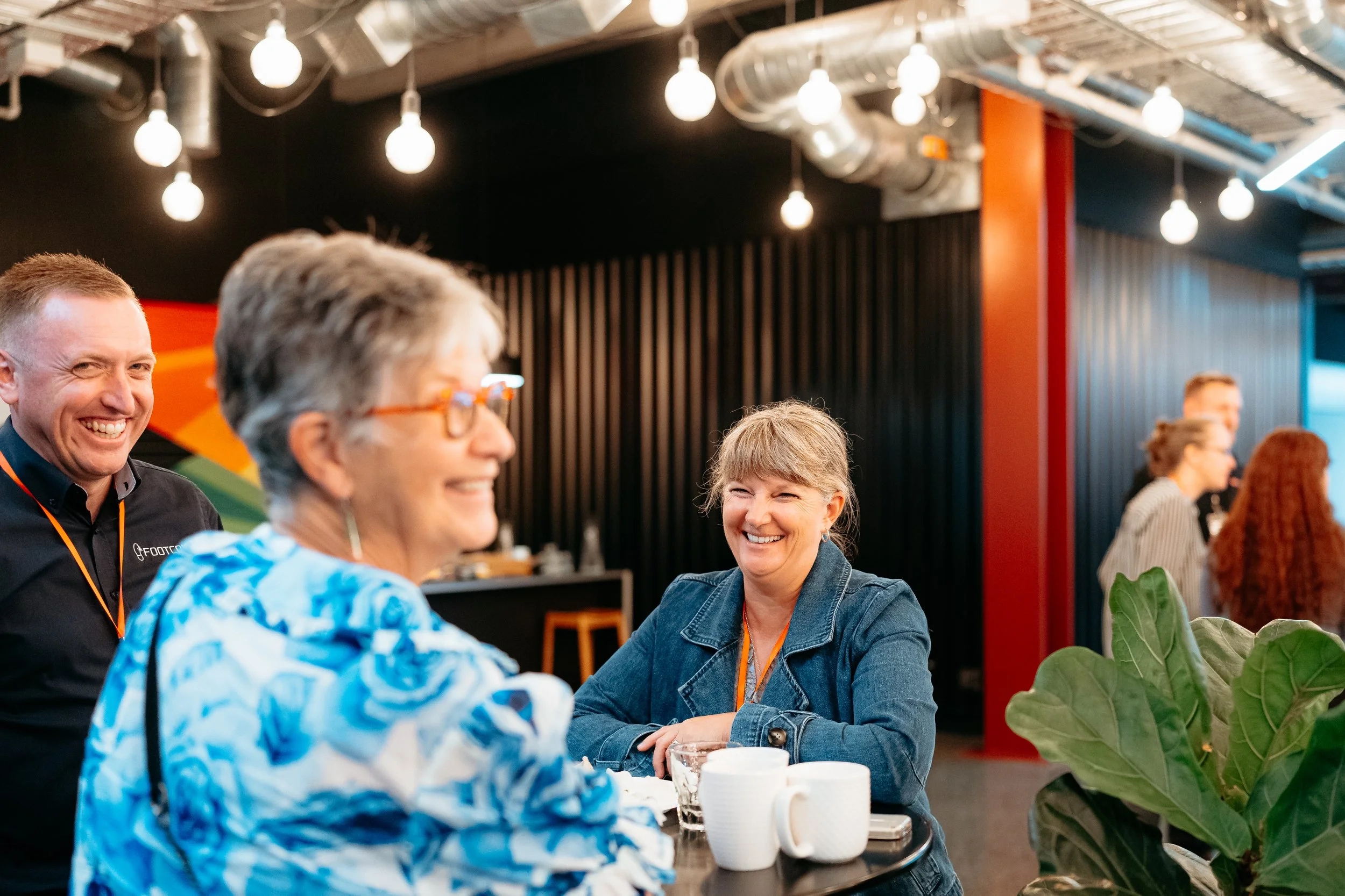 People smiling and chatting at a bar or cafe counter in a modern indoor setting with hanging lights and black wall panels.