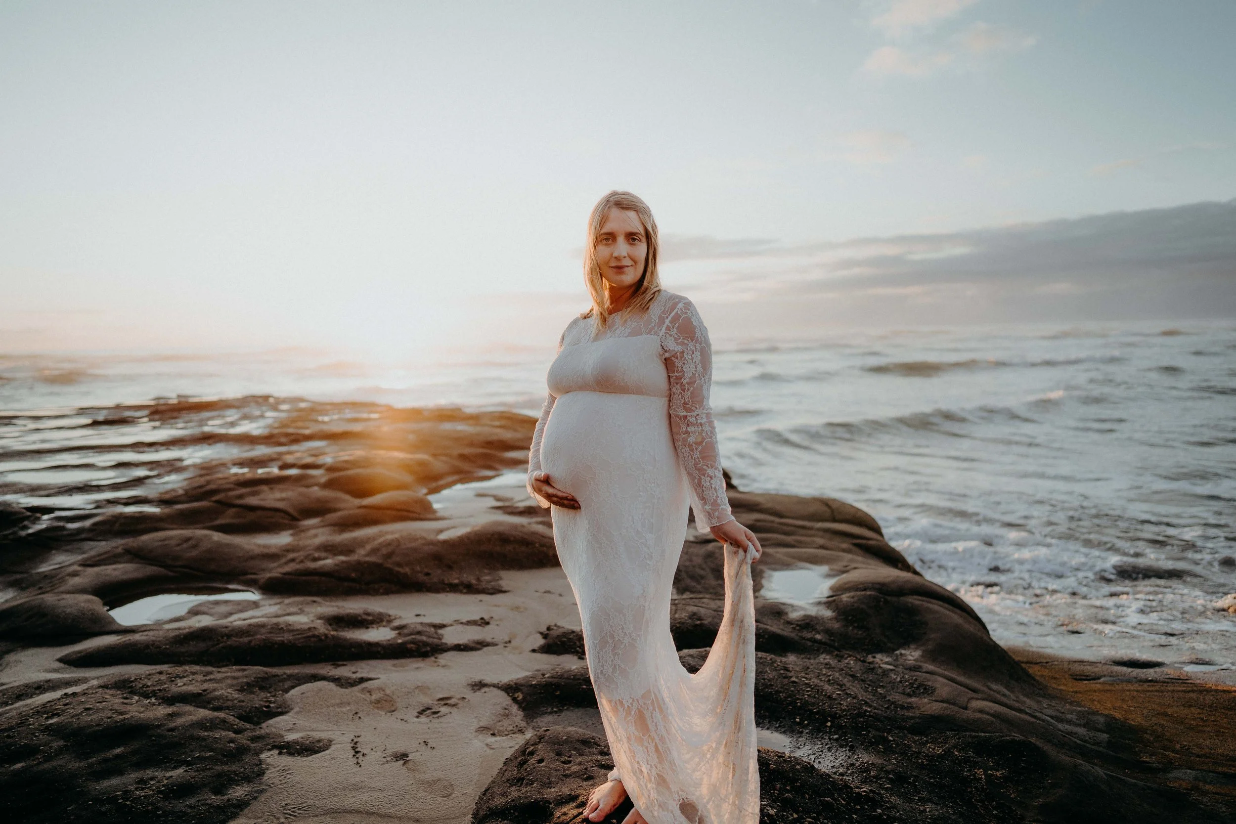 A pregnant woman in a white lace dress standing on rocks at the beach during sunset.