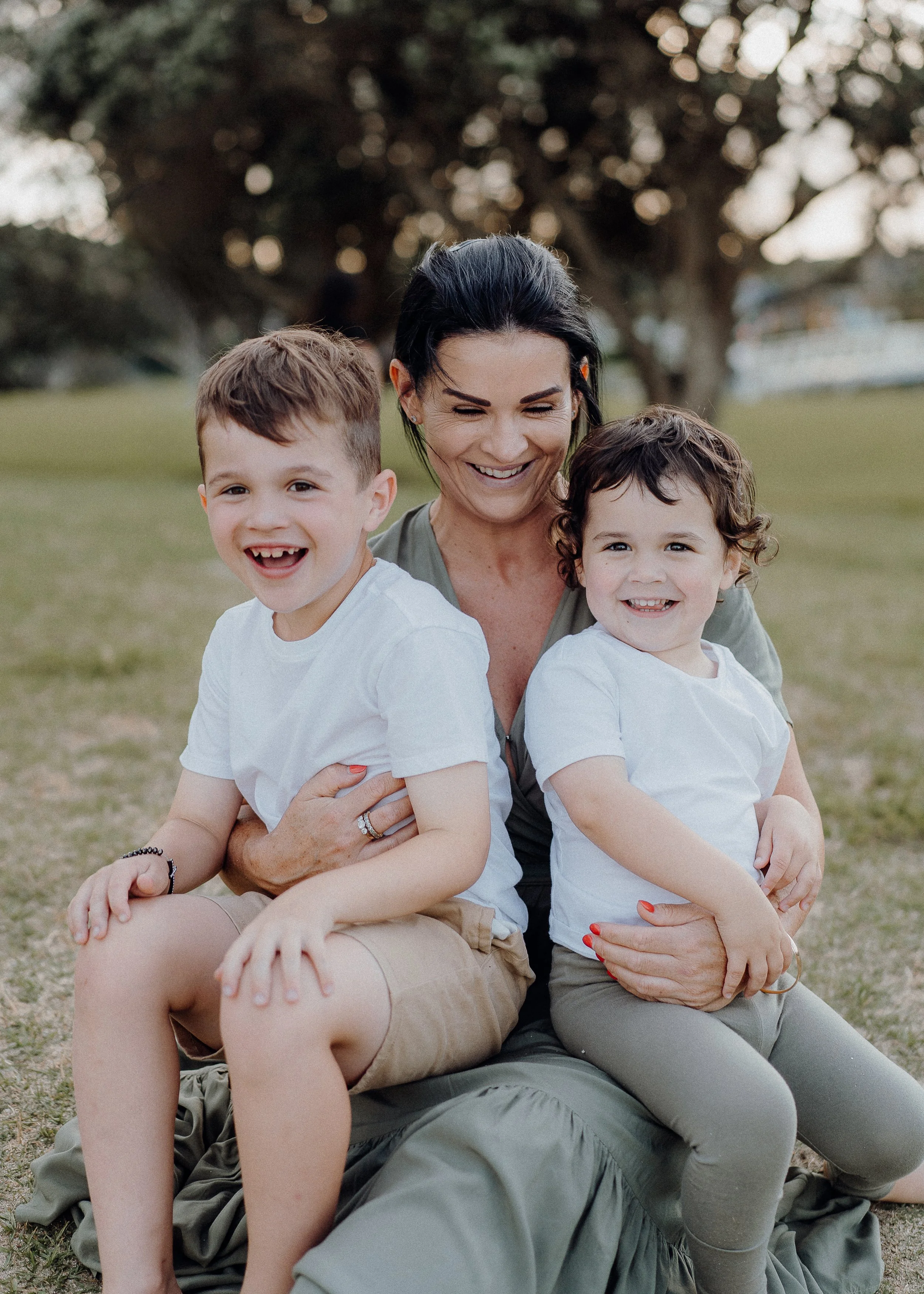 A woman sitting on the ground outdoors with two children, all smiling and laughing. Two young kids are sitting on her lap, wearing white shirts, in a park setting with grass and trees in the background.