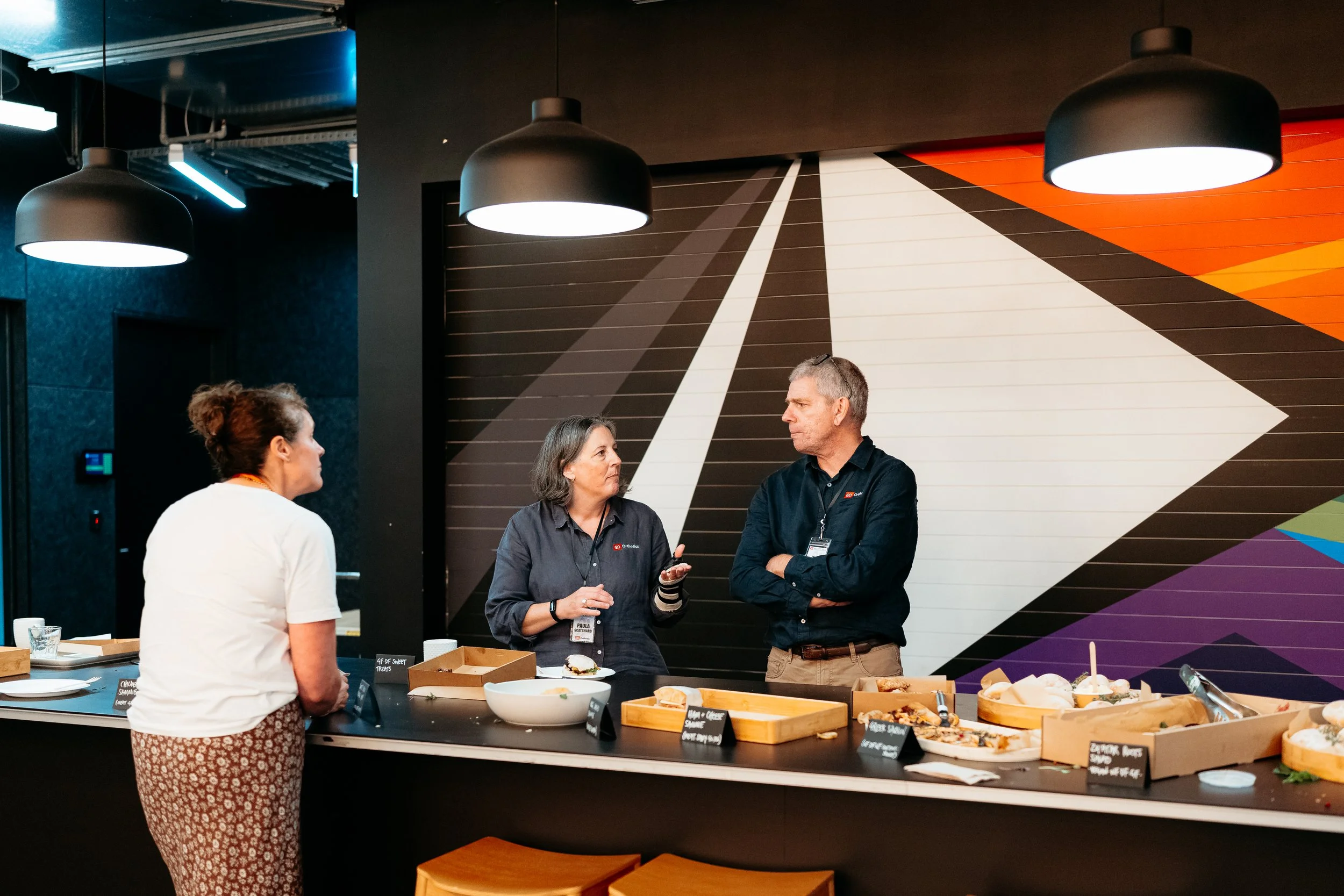 Three people talking at a buffet table in a modern room with black pendant lights, a colorful abstract mural, and assorted food on trays.