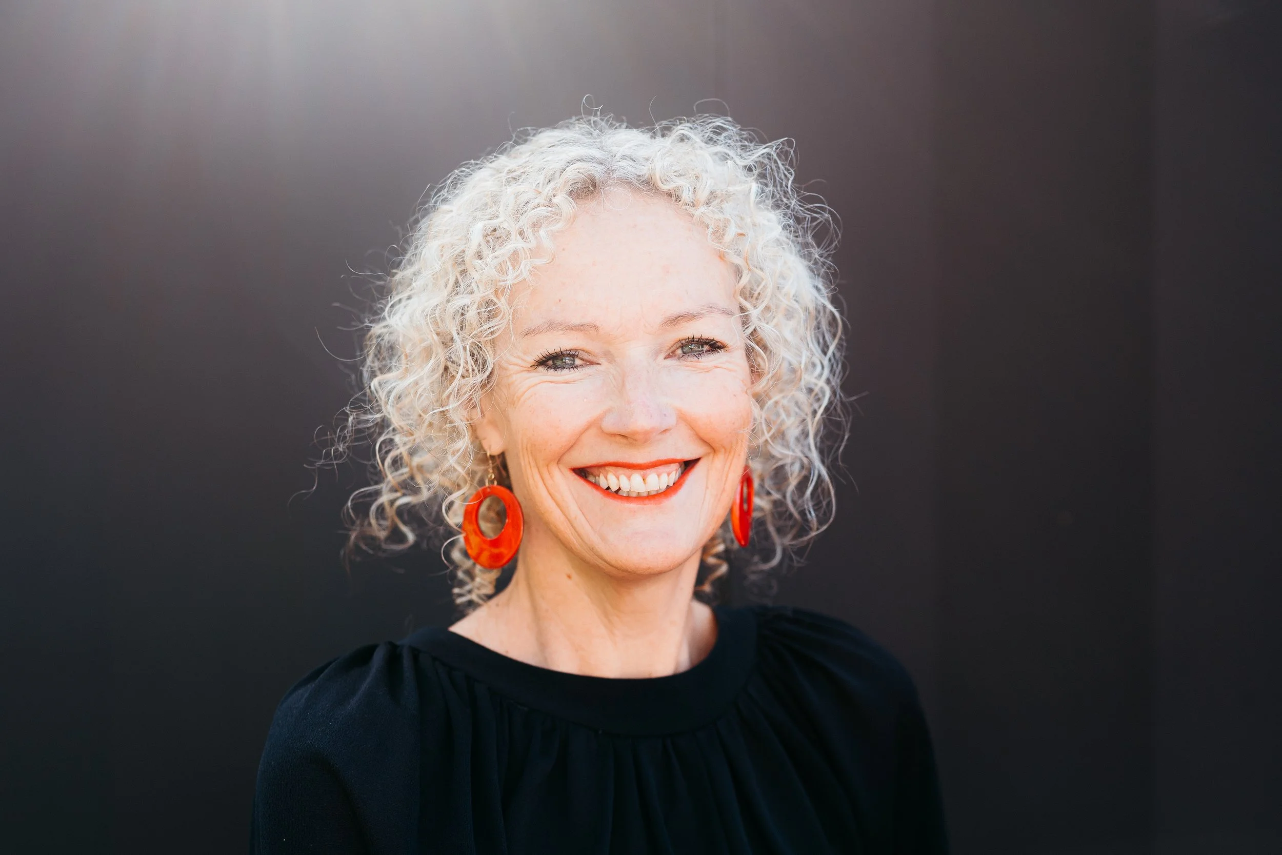 A smiling woman with curly gray hair, wearing red earrings and a black top, stands against a dark background.