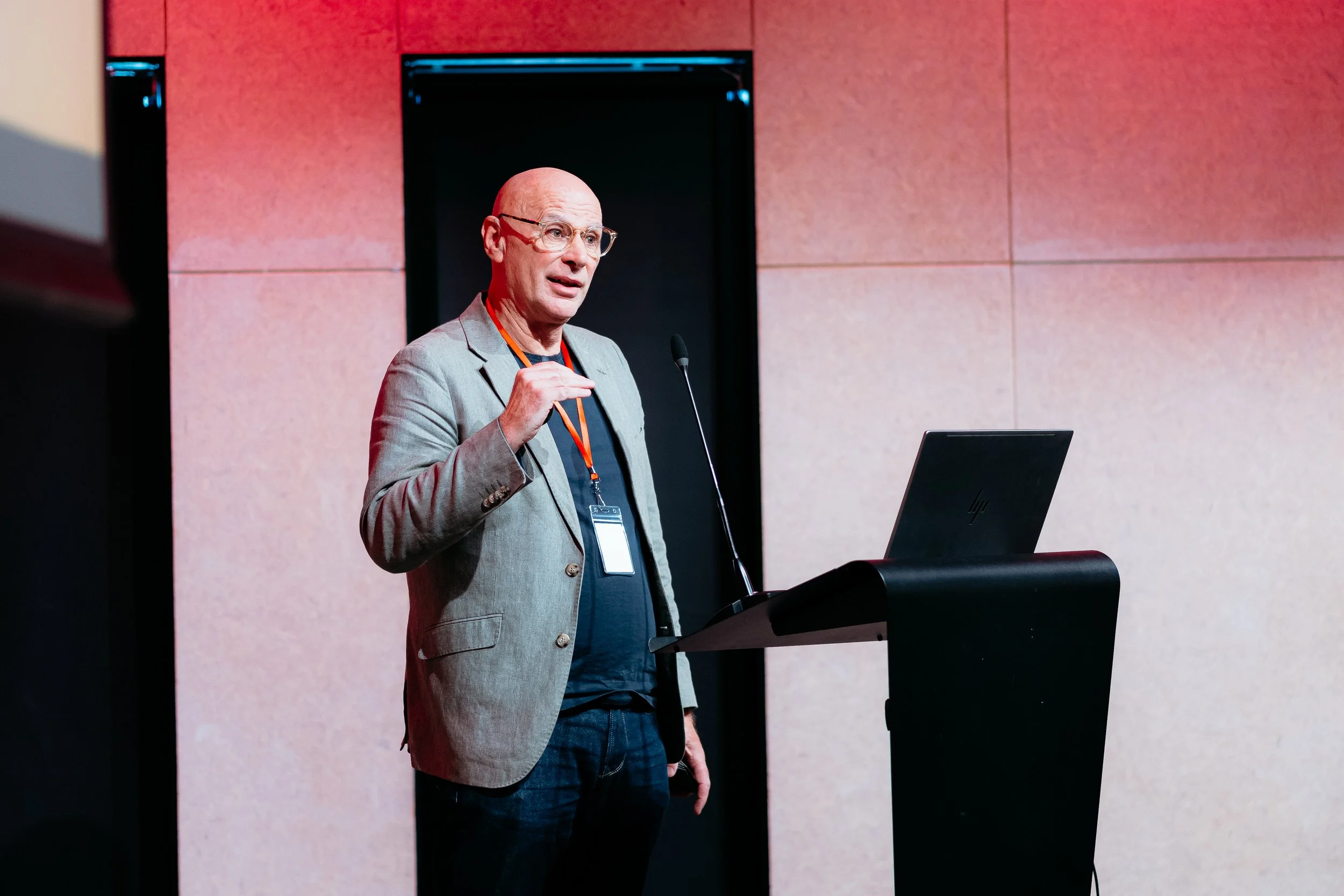 A man with glasses and a bald head giving a presentation at a conference, standing behind a podium with a laptop, wearing a gray blazer, dark shirt, and an orange lanyard.