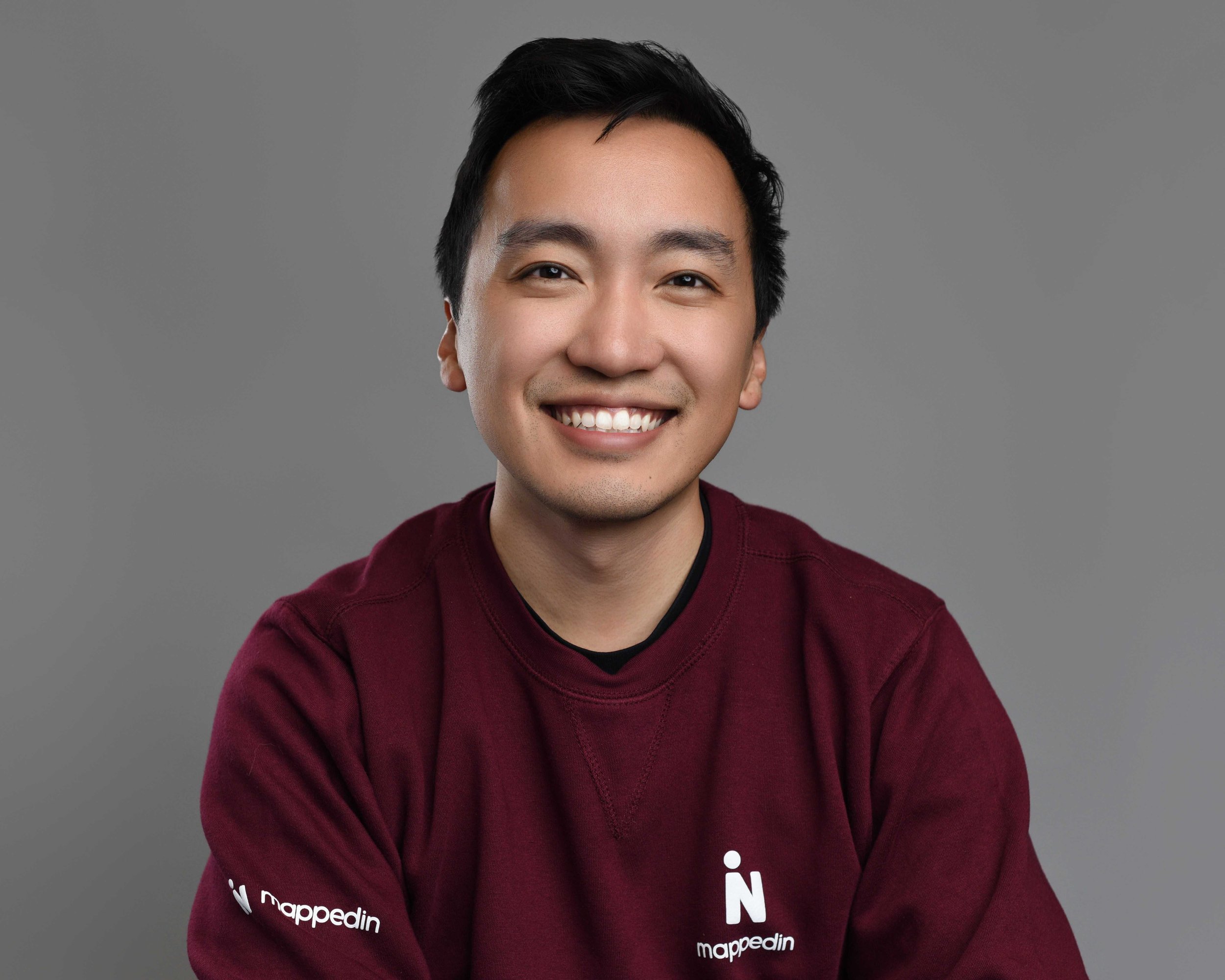 Smiling young man with short black hair wearing a maroon sweater with the word "mappedin" written on it, set against a plain gray background.