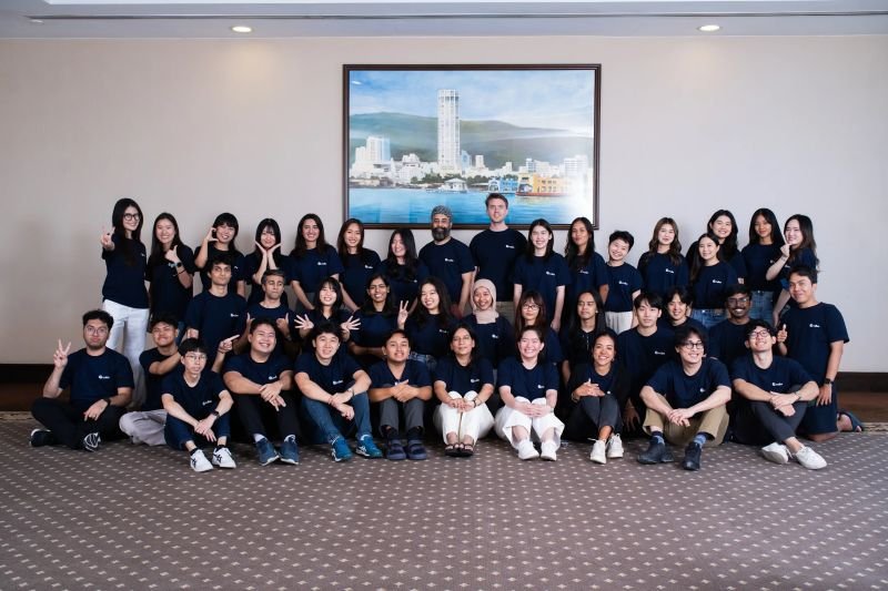 Group of young adults and middle-aged people in matching navy blue shirts posing for a group photo in a conference room with a cityscape painting in the background.
