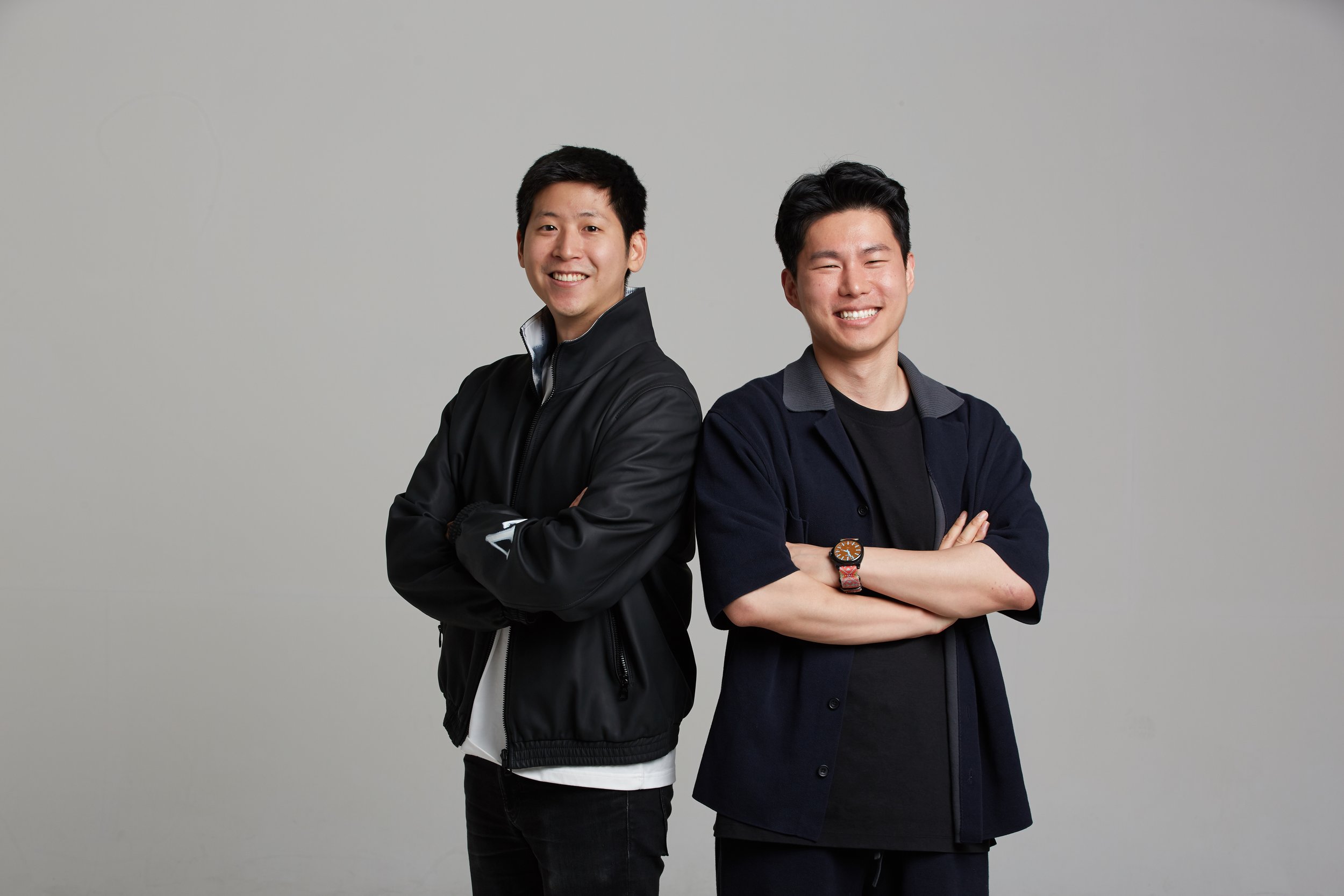 Two young men standing with arms crossed, smiling, against a plain gray background.