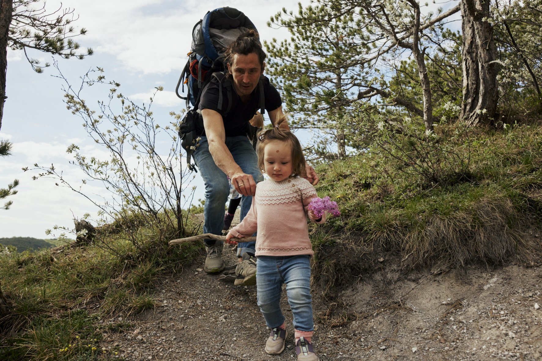 Hier im Wald fühlt sich Anna sichtlich wohl und hat sich nun einen steilen, erdigen Pfad rauf zum Jubiläumskreuz vorgenommen. „Lass den Stock liegen, wenn du da raufgehst“, ruft Bernhard ihr nach. Aber das wird nichts, also muss ihr Papa Anna mit beim Aufstieg mit den vollen Händen helfen. Beim Kreuz angekommen, gibt es für unsere kleinen Bergführerinnen eine Pause samt Stärkung. Danach führt unser Weg wieder Richtung Hütte. Die Mädchen verstecken sich hinter Bäumen und Büschen, riechen an blühenden Felsenbirnensträuchern, inspizieren Nadeln kleiner Föhren und Baumkeimlinge neben dem Weg. Sie lassen sich treiben. Und wir auch. - 