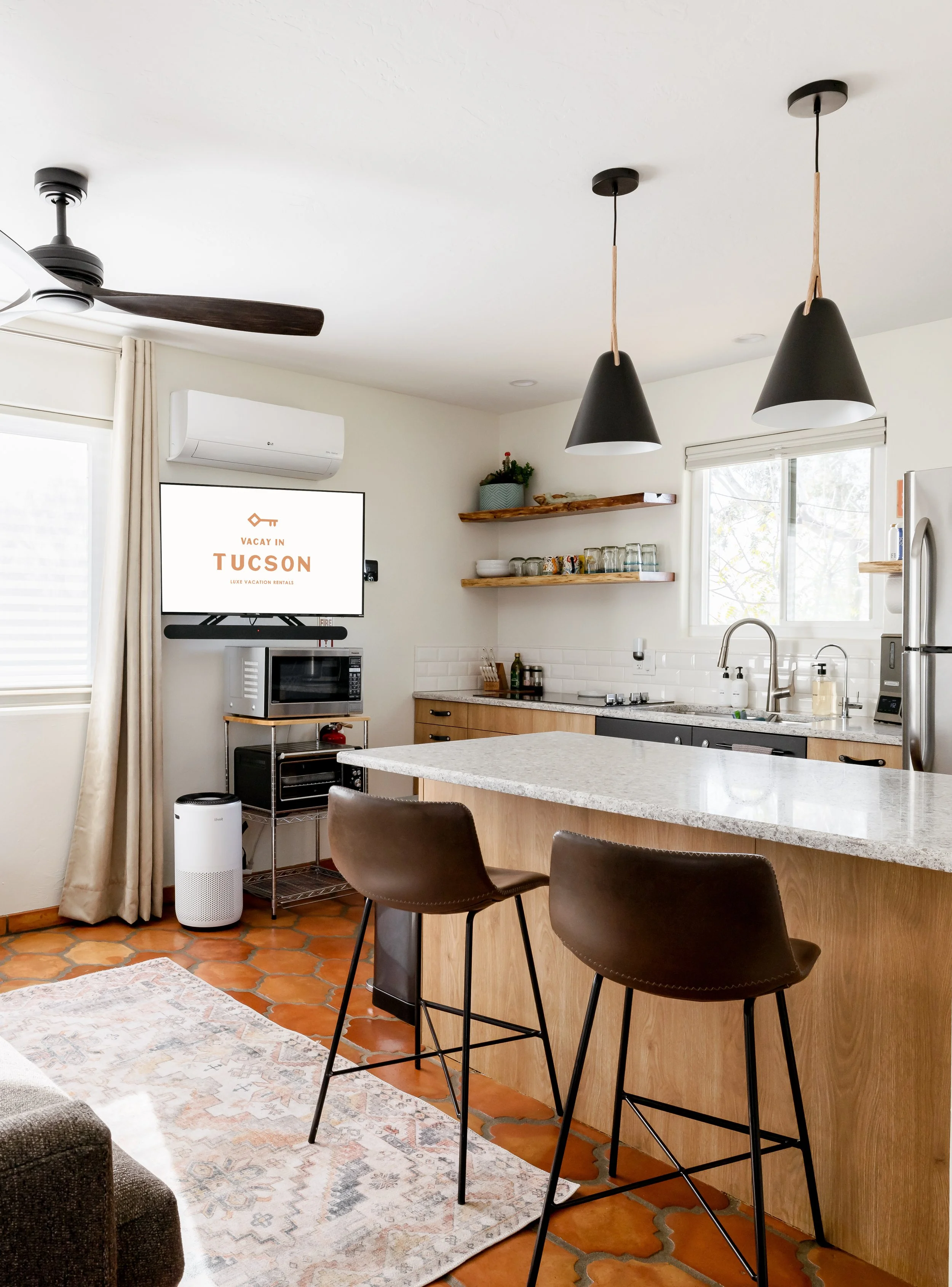 White kitchen with a stone peninsula, two brown bar stools, black pendant lights, open wood shelving, terracotta tile flooring, and a television visible beyond the island.