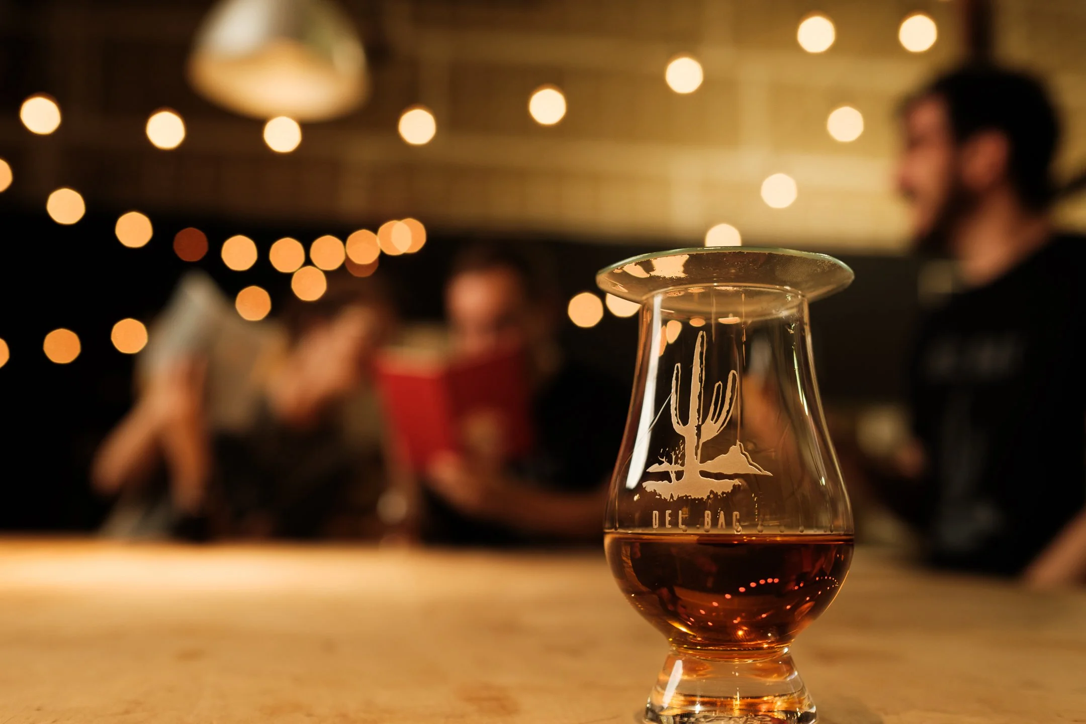Glass of amber whiskey etched with a saguaro design sits on a wooden counter with blurred people and warm string lights in the background.