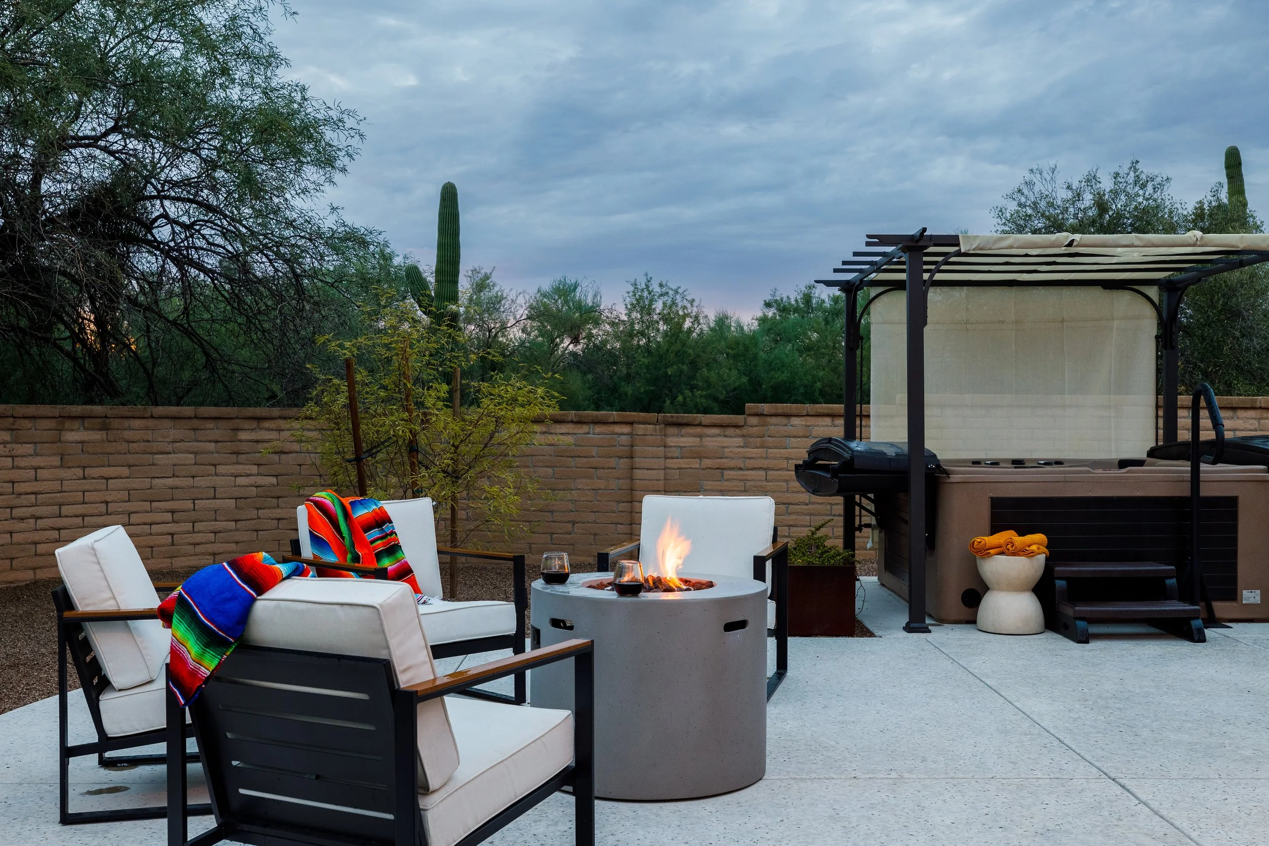 White cushioned patio chairs with colorful striped blankets surround a glowing round fire pit beside a covered hot tub and desert landscaping.