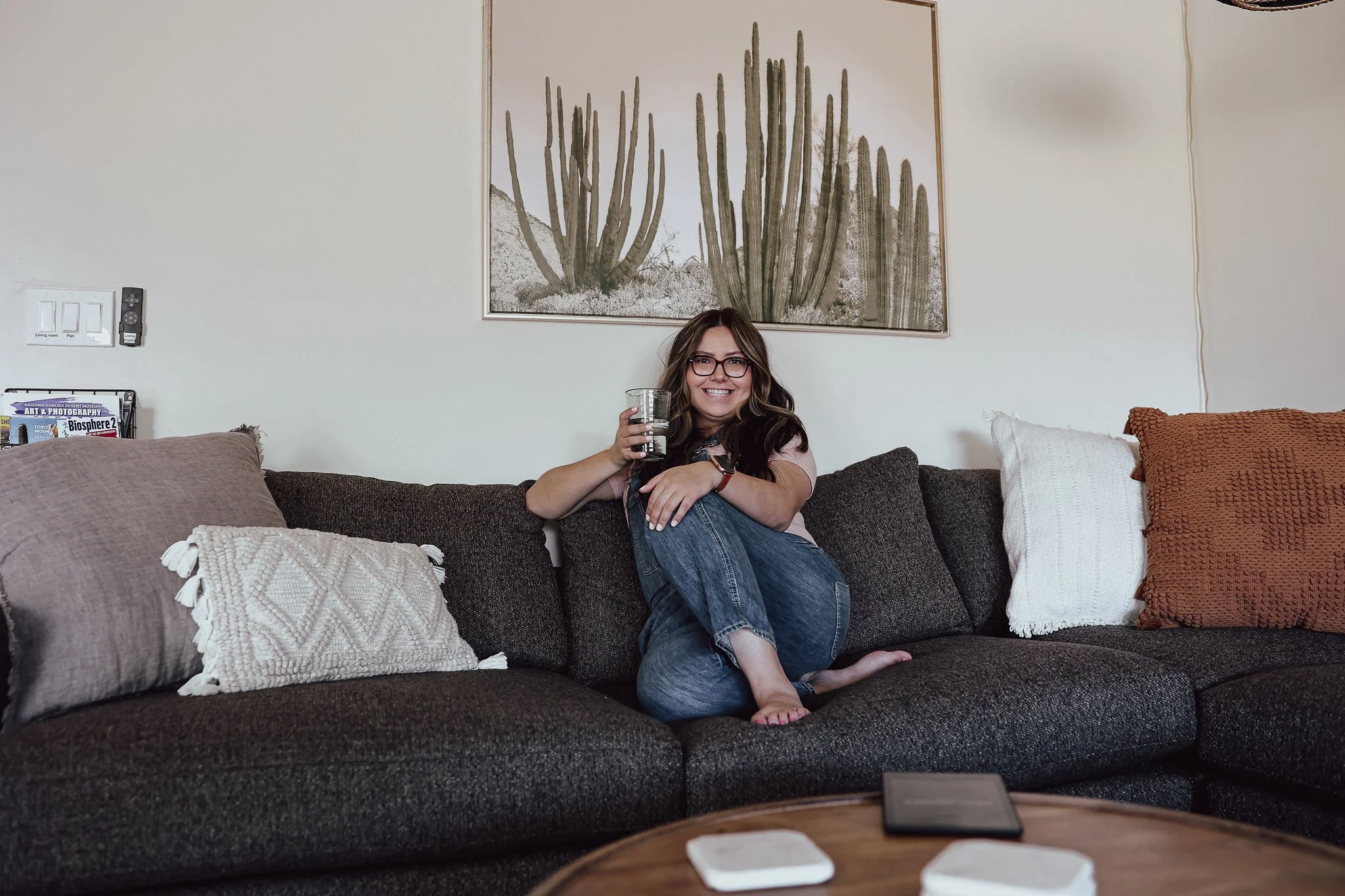 Woman sits cross-legged on a dark sectional sofa at The Verde House, smiling with a drink in hand beneath framed cactus artwork and surrounded by neutral textured pillows.