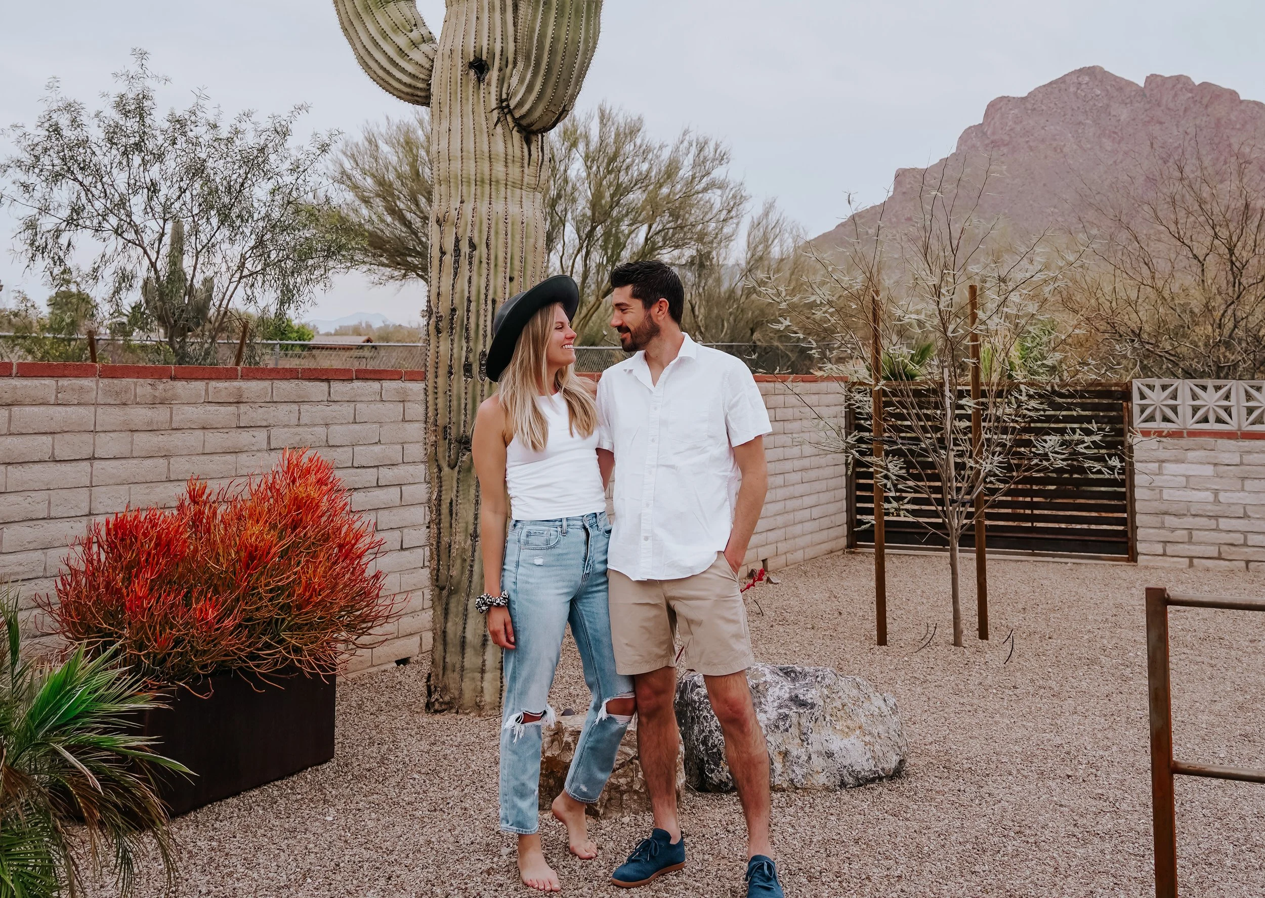 Smiling couple stands together beside a tall saguaro in landscaped backyard, with desert plants and rugged mountains in the background.