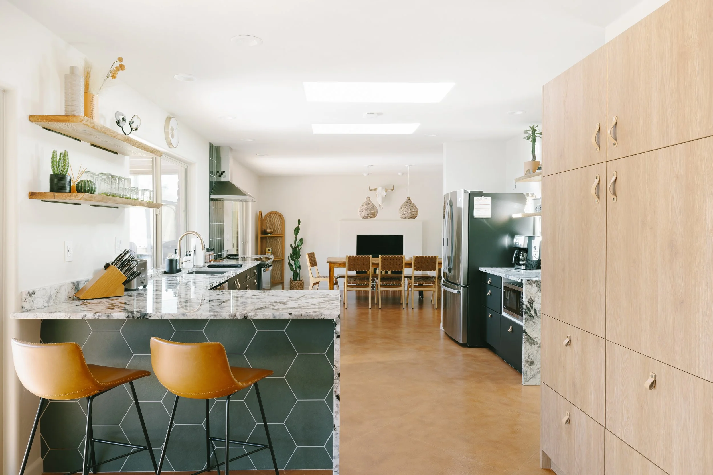 Open kitchen with marble-patterned counters, dark cabinetry, wood shelves, two brown counter stools, and a dining table visible in the adjoining room.