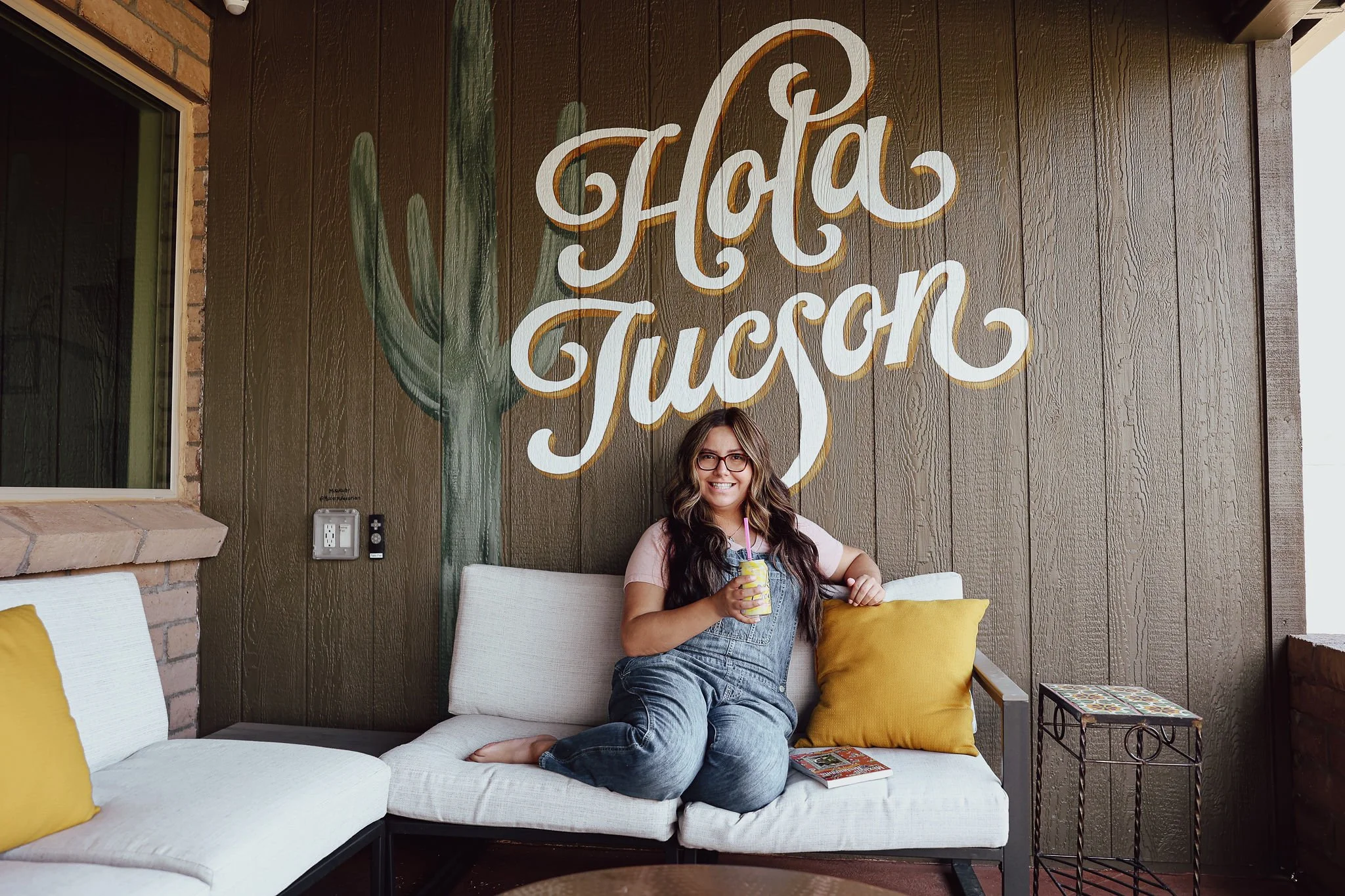 Woman with long brown hair and glasses sits on a cream patio sofa holding a drink beneath a large hand-painted "Hola Tucson" mural and saguaro.