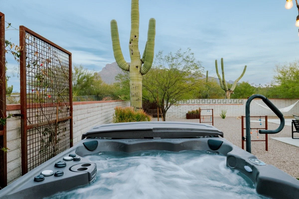 A bubbling hot tub in front of a towering saguaro cactus and Catalina Mountain views beyond a low desert wall, with native landscaping surrounding the patio.
