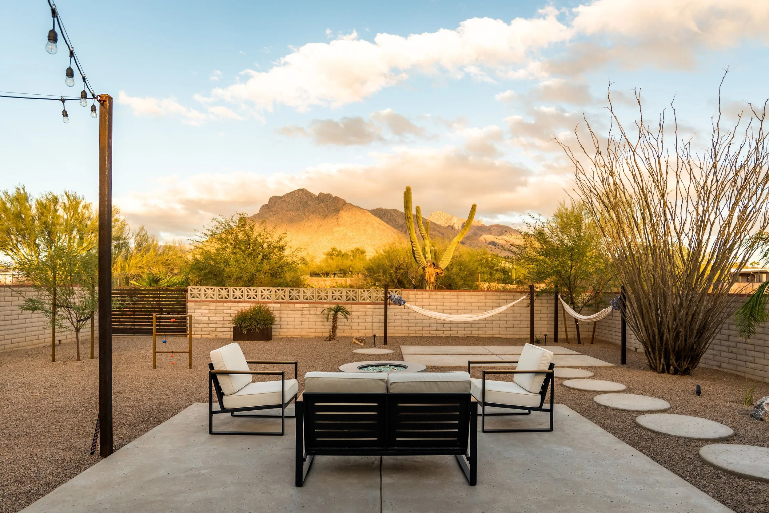 Backyard with two white hammocks along a low brick wall, cushioned patio seating around a fire pit, a tall saguaro, desert plants, and mountains in the background.