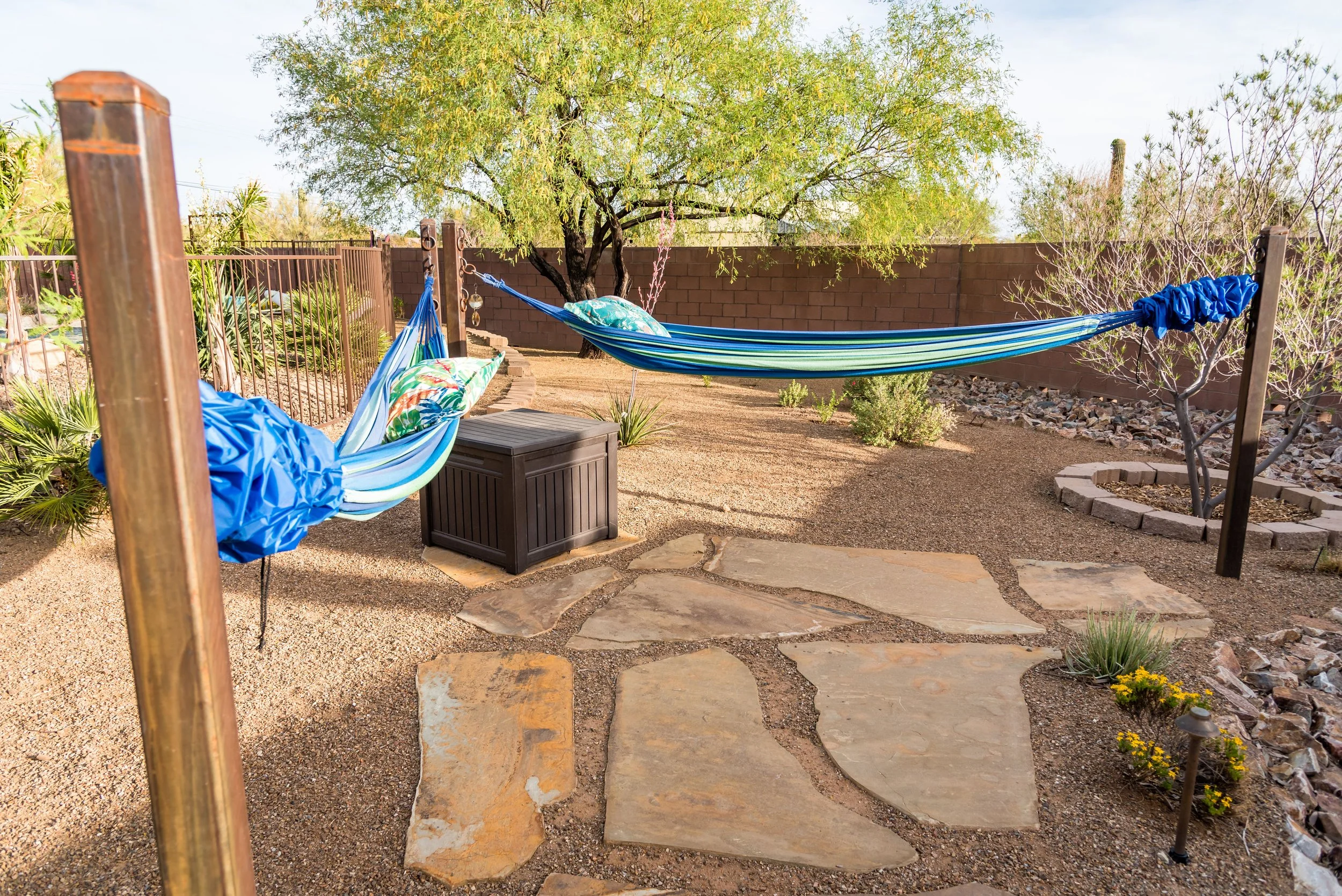 Spacious desert backyard with two hammocks stretched between posts, gravel landscaping, mature trees, and surrounding greenery.