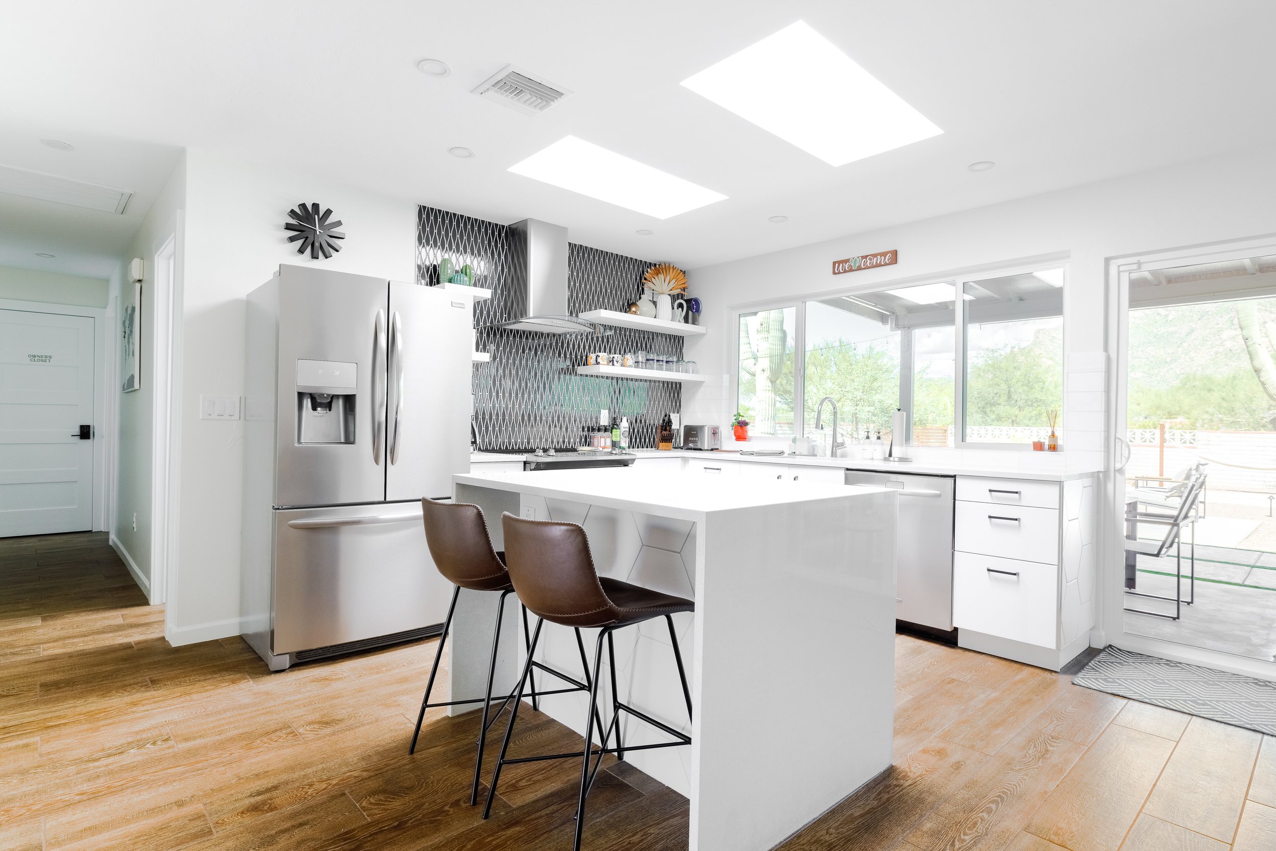 Modern white kitchen at Casa Palma with stainless-steel appliances, a large island with brown bar stools, open shelving, patterned backsplash, skylights, and windows overlooking the backyard.