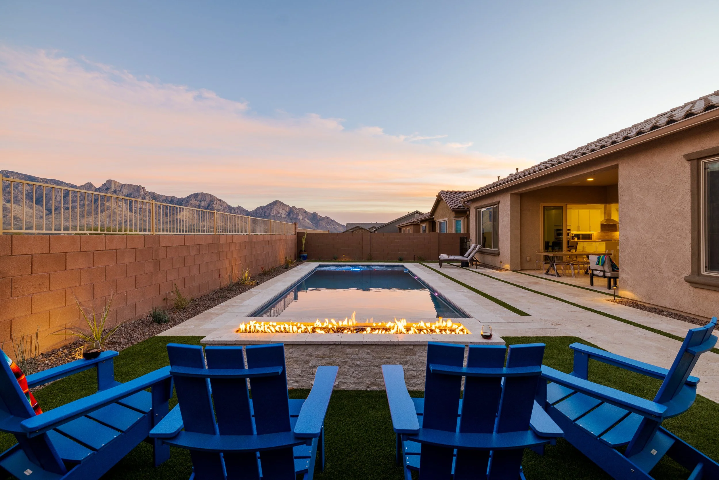 Four bright blue Adirondack chairs face a long rectangular pool and glowing linear fire feature with mountains beneath a pastel evening sky.