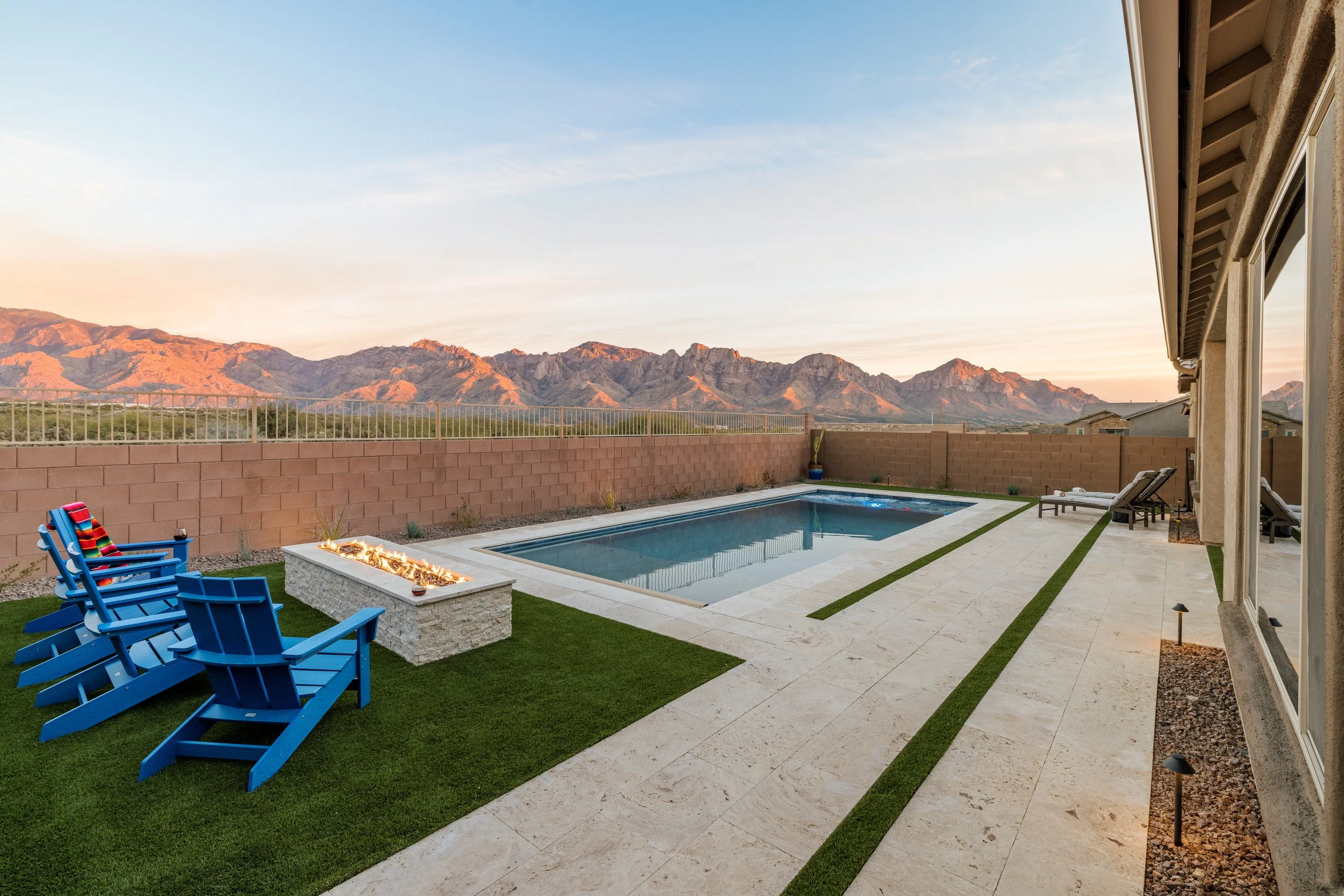 Rectangular pool beside a glowing linear fire feature and blue Adirondack chairs, with rugged mountains lit pink by the setting sun.