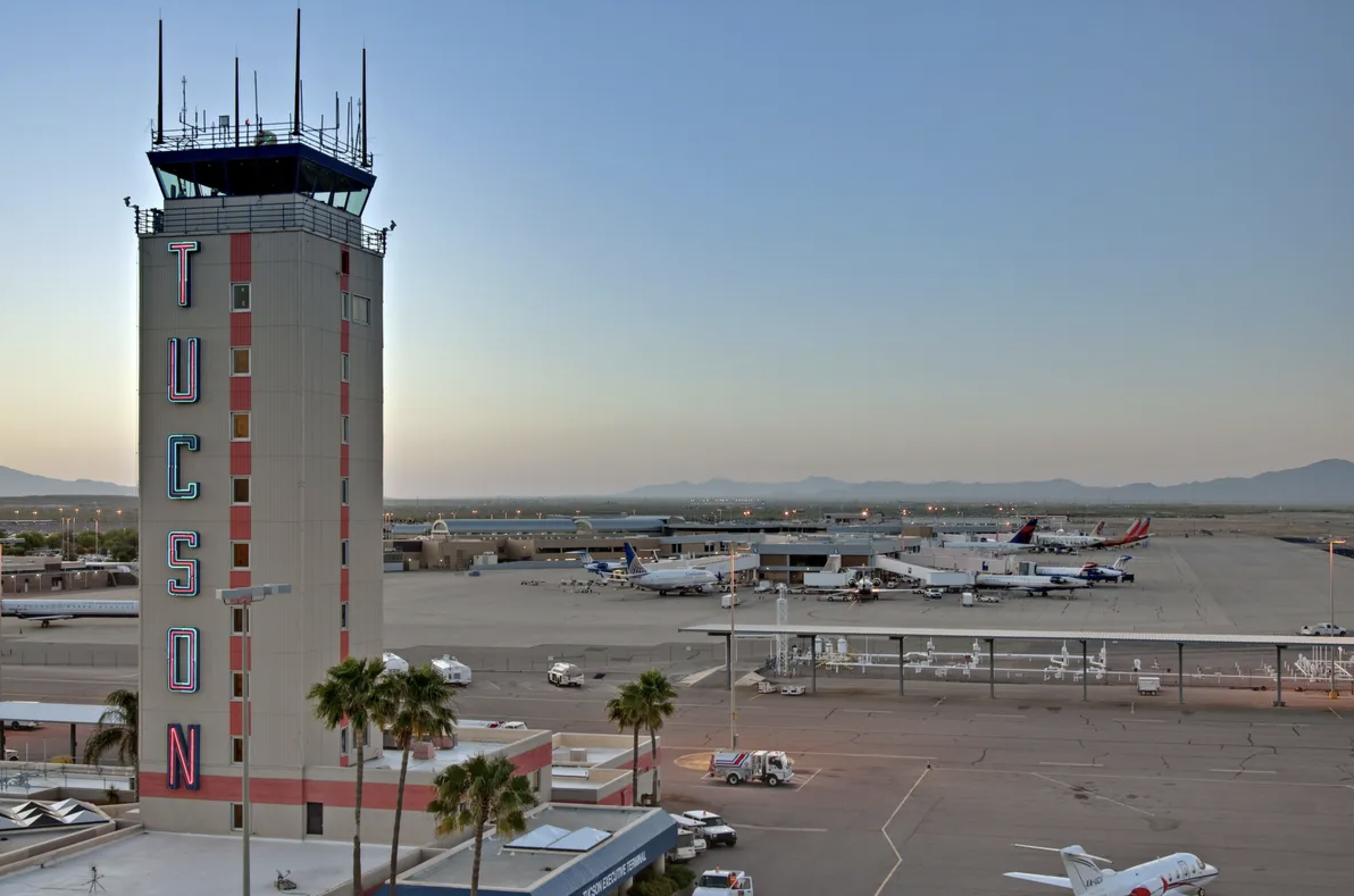 Airport control tower with large vertical letters spelling TUCSON stands beside runways, parked airplanes, palm trees, and distant mountains.