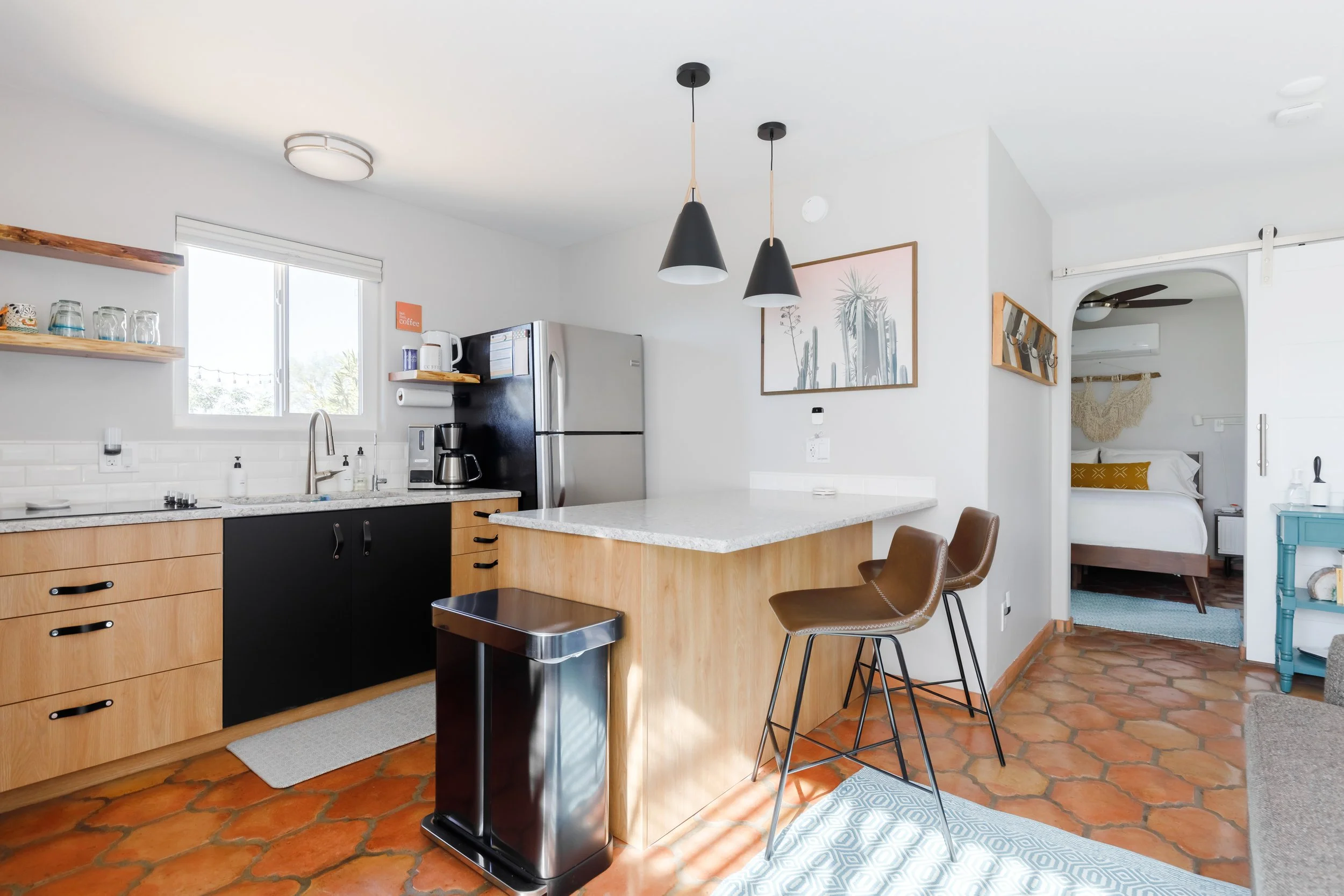 Bright kitchen with light wood and black cabinets, stainless steel appliances, white countertops, two brown bar stools, and a bed visible through an arched doorway.