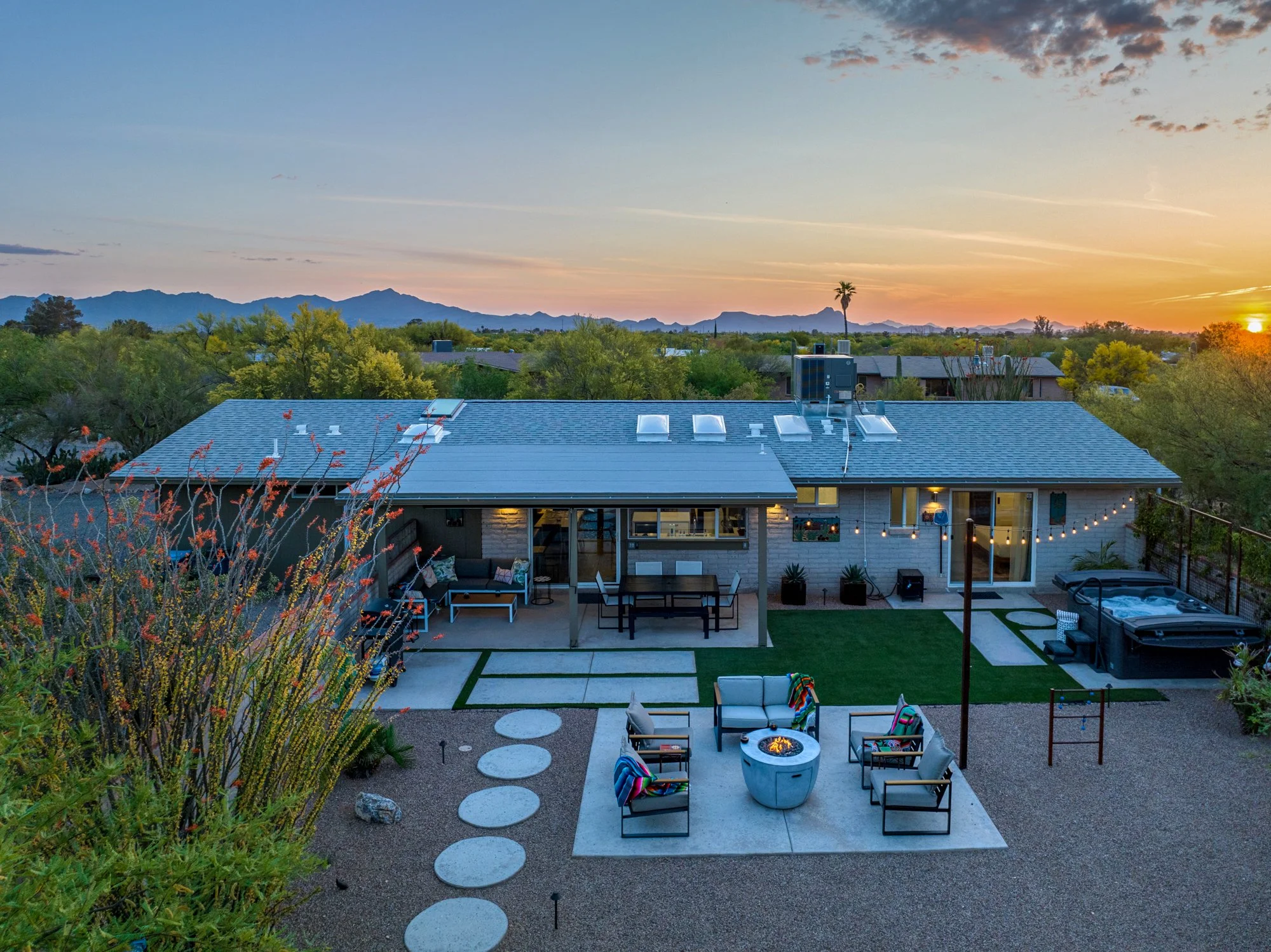 Elevated sunset view over Casa Palma’s backyard with a fire pit seating area, green lawn, desert landscaping, hot tub, string lights, and distant mountain silhouettes.