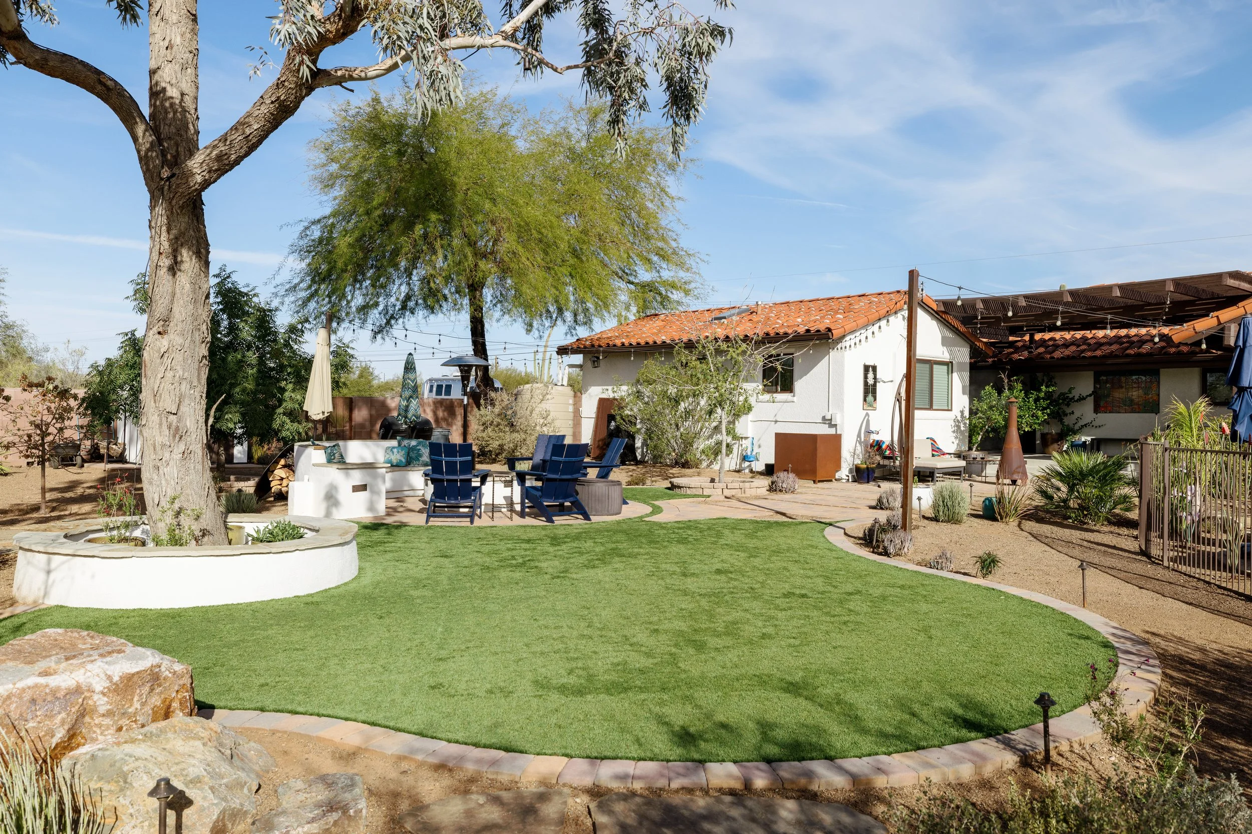 Spacious backyard with artificial grass, mature trees, blue Adirondack chairs, a circular fire pit, patio seating, desert landscaping, and a white stucco home with a red tile roof.