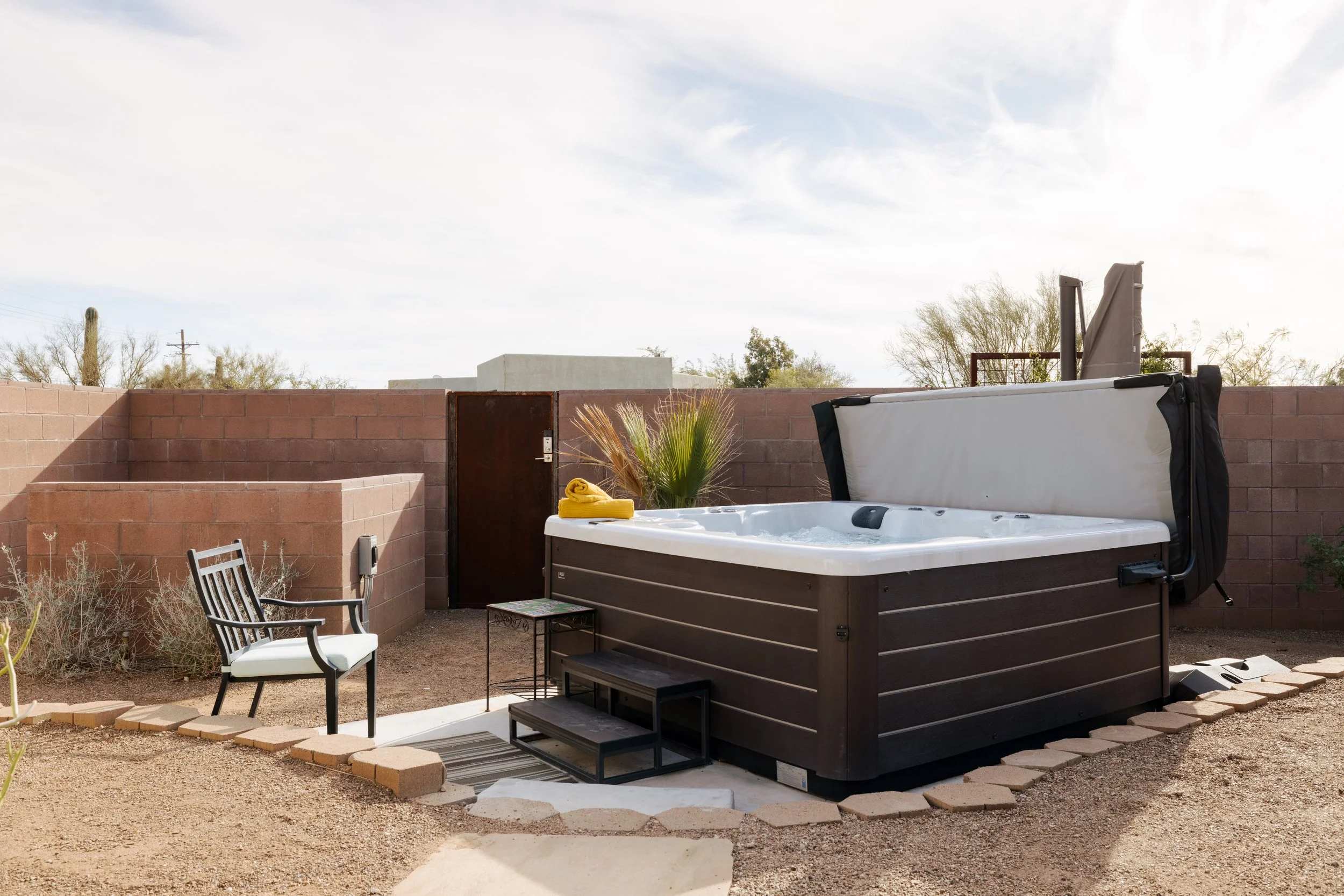 Large square hot tub with a raised cover, steps, a small side table, single patio chair, gravel ground, and low block walls around the yard.