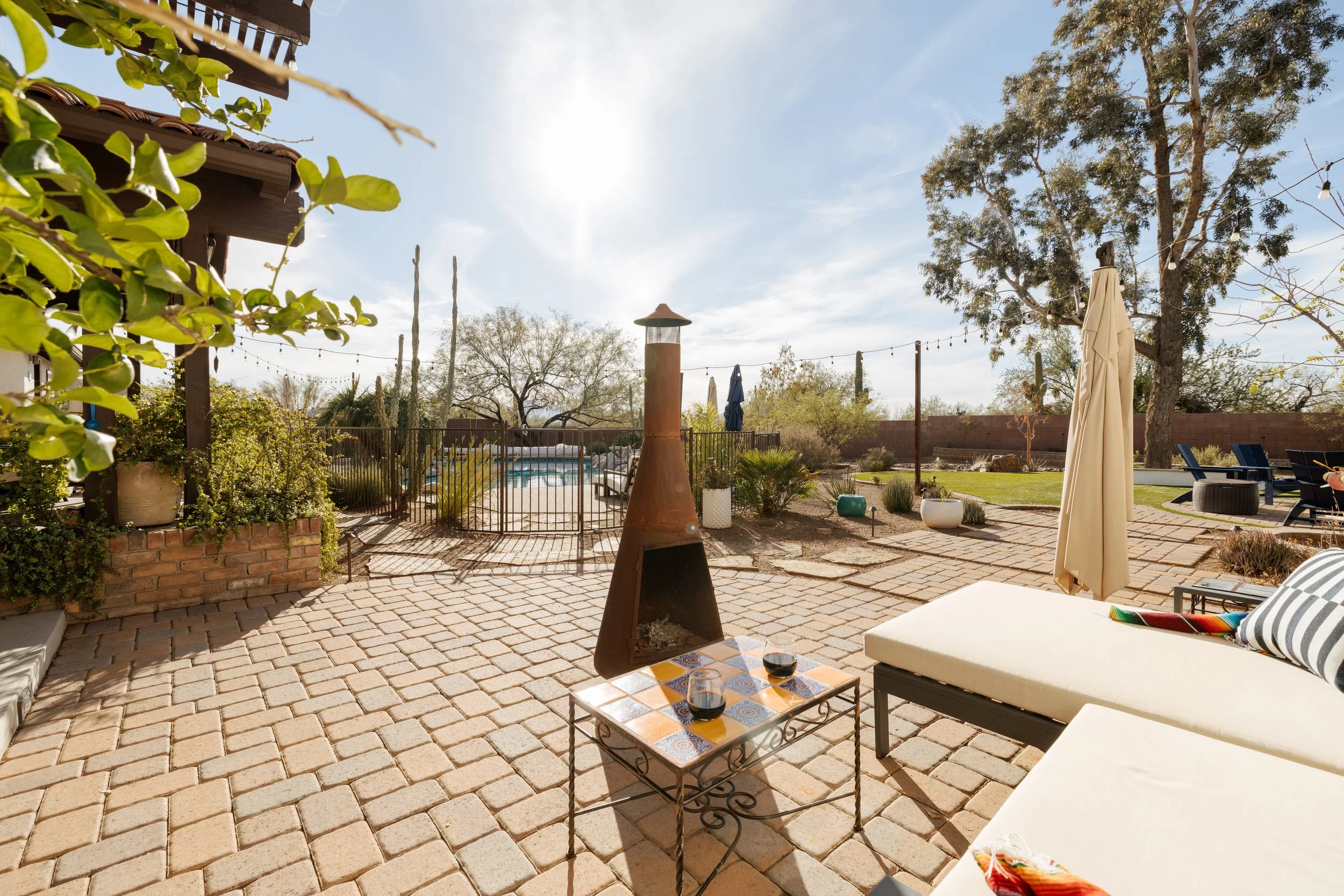 Sunny brick patio with a cushioned outdoor sectional, tiled coffee table, tall metal chiminea, string lights, umbrellas, and a fenced swimming pool beyond.