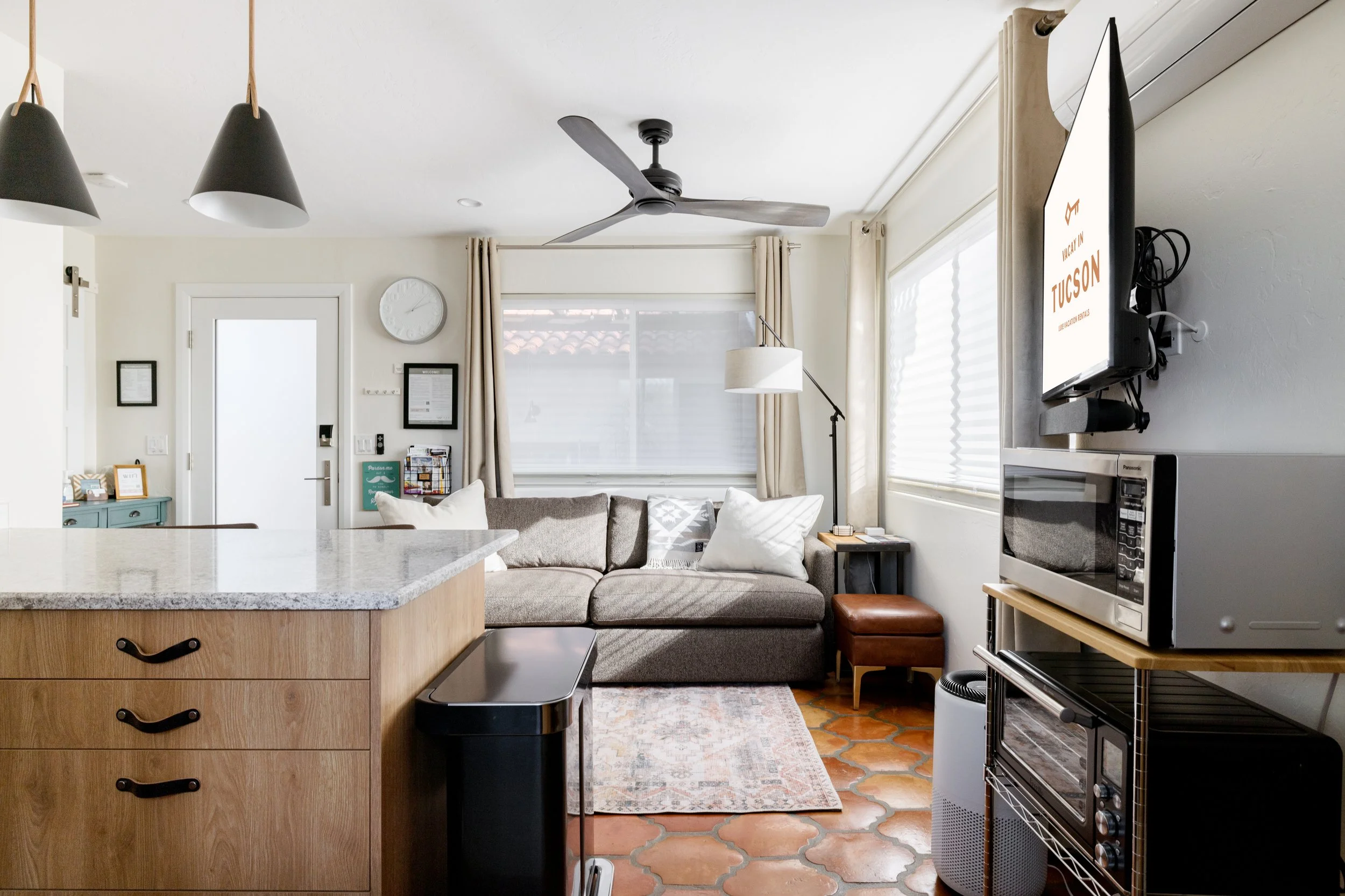Open living area with a gray sectional sofa, patterned rug, light wood kitchen cabinets, large windows, microwave shelving, and a wall-mounted television.