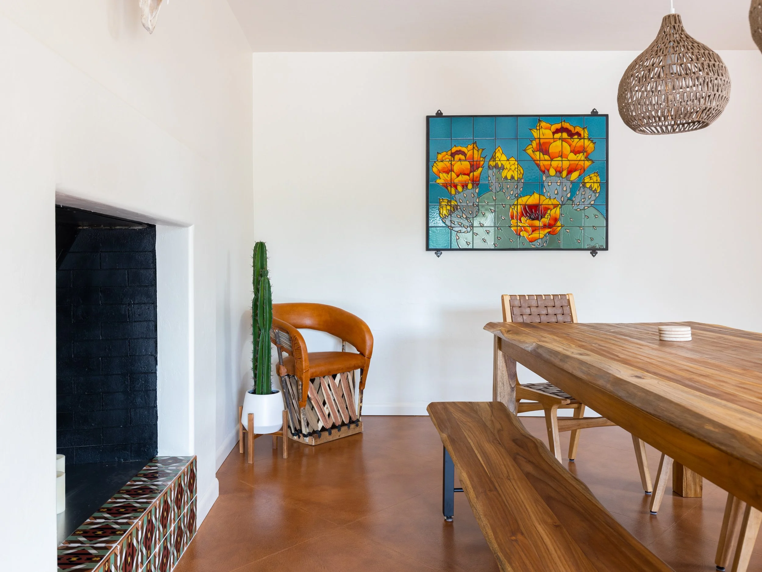 Wood dining table and bench beside a black fireplace with a colorful tiled hearth and a large orange-flowering cactus tile mural on the wall.