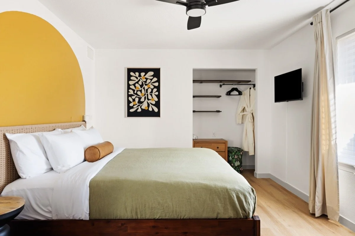Bed with pale green bedding and woven headboard set against a large mustard-yellow painted arch, with floral artwork and an open closet.