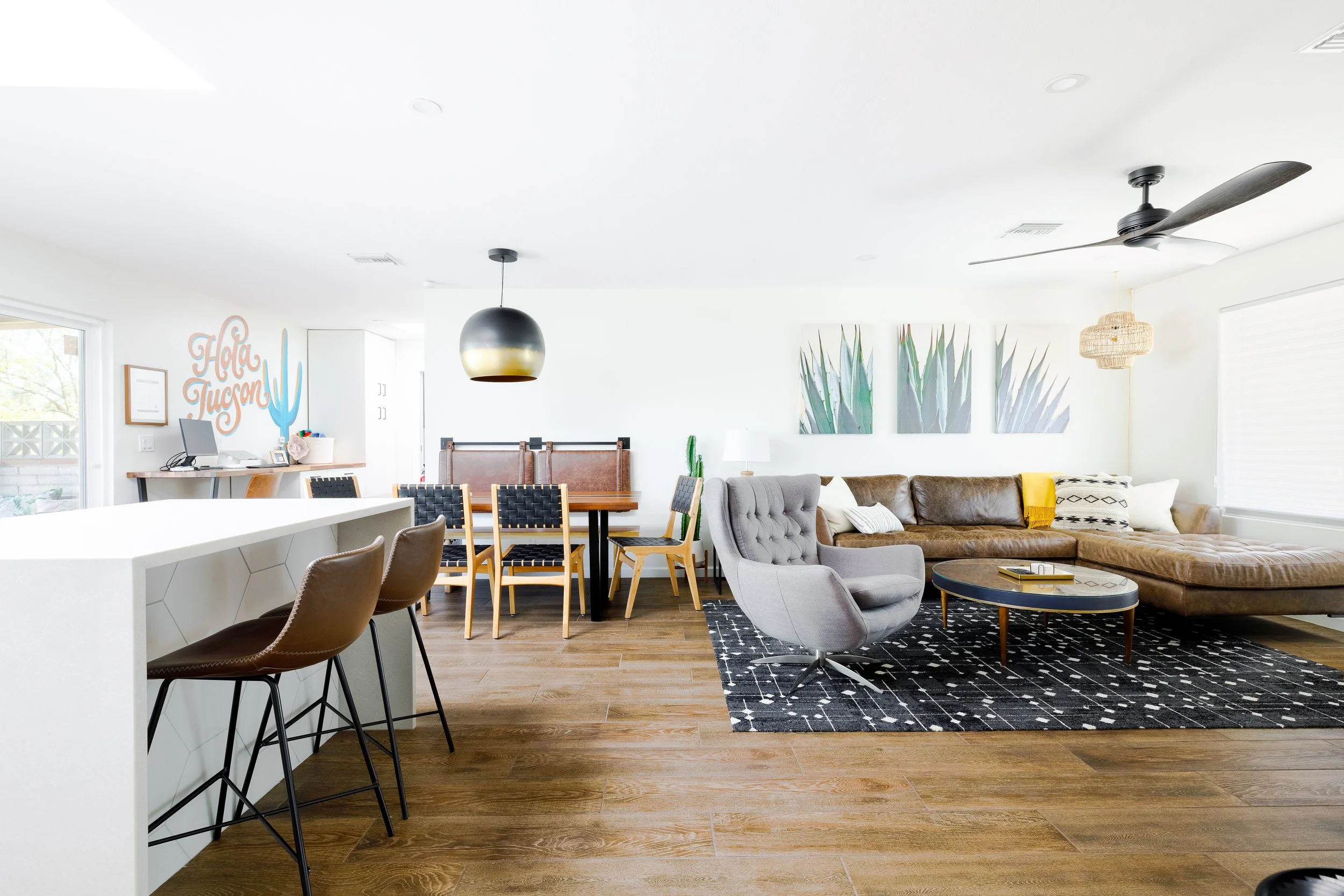 Spacious open living area at Casa Palma with a sectional leather sofa, gray lounge chair, dining table, kitchen island seating, modern desert décor, and a colorful Hola Tucson mural on the far wall.