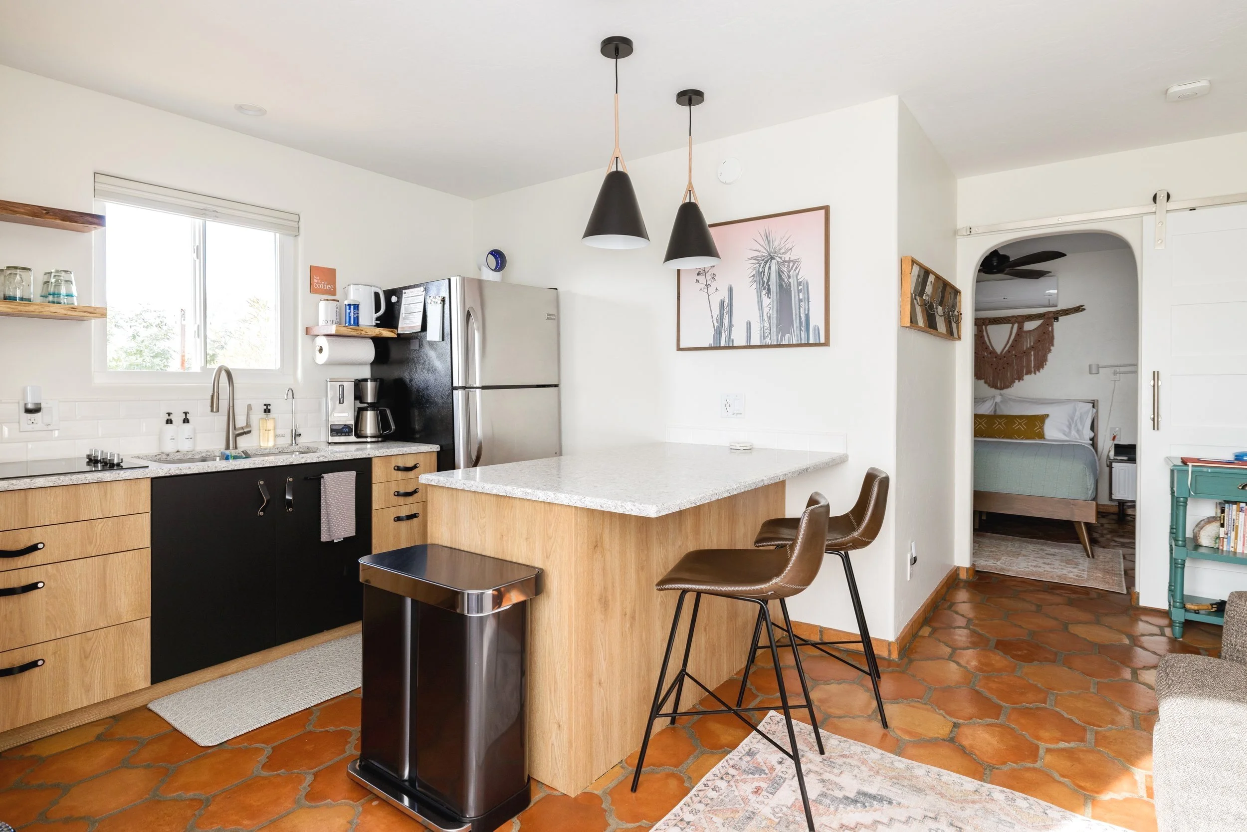 Kitchen with light wood and black cabinets, stainless steel refrigerator, white countertops, two brown bar stools, pendant lights, and a bedroom visible through an arched doorway.